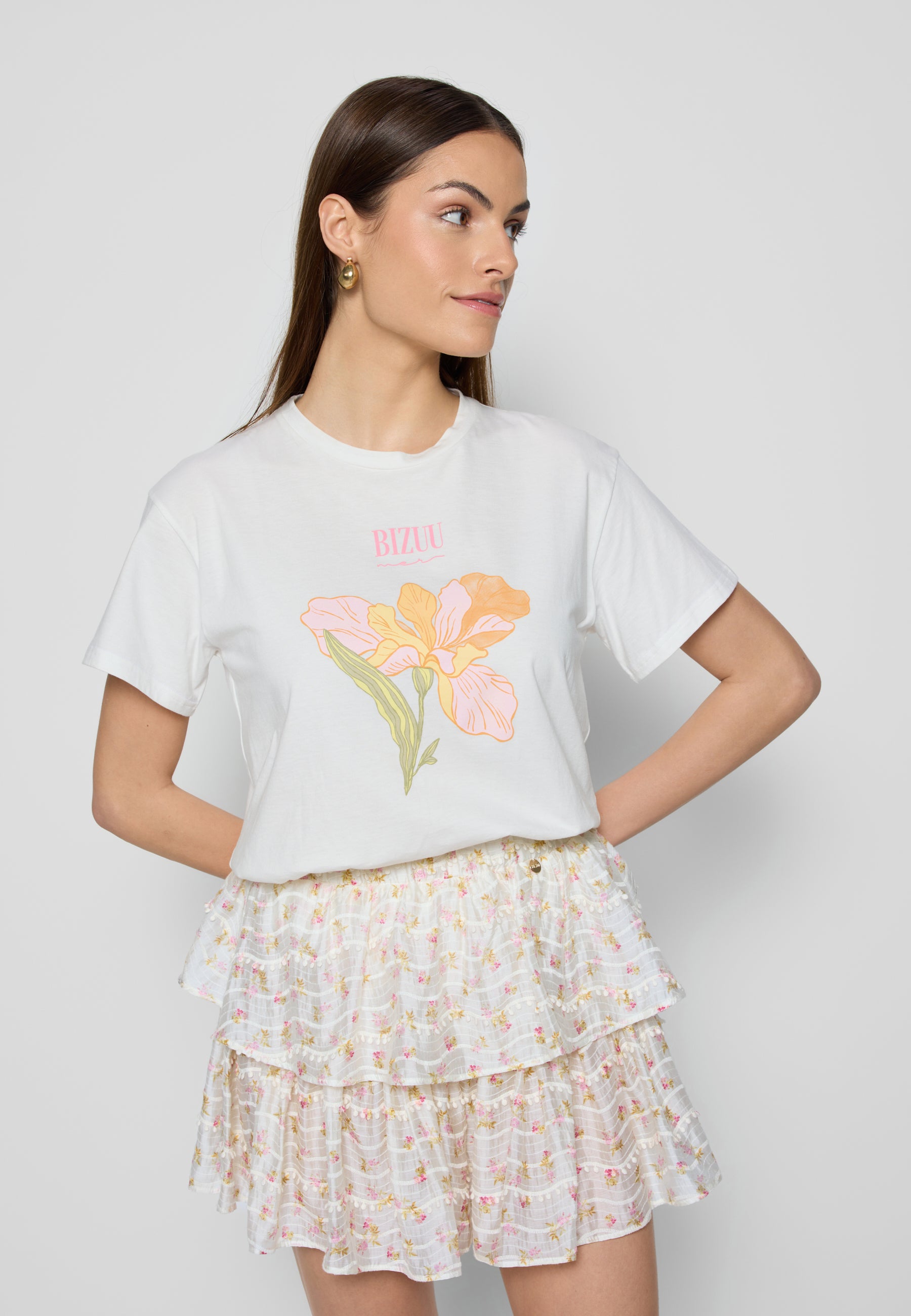 Modelka w białym t-shircie z kwiatowym nadrukiem i białej spódniczce w kwiatki z falbanami, widok z profilu.