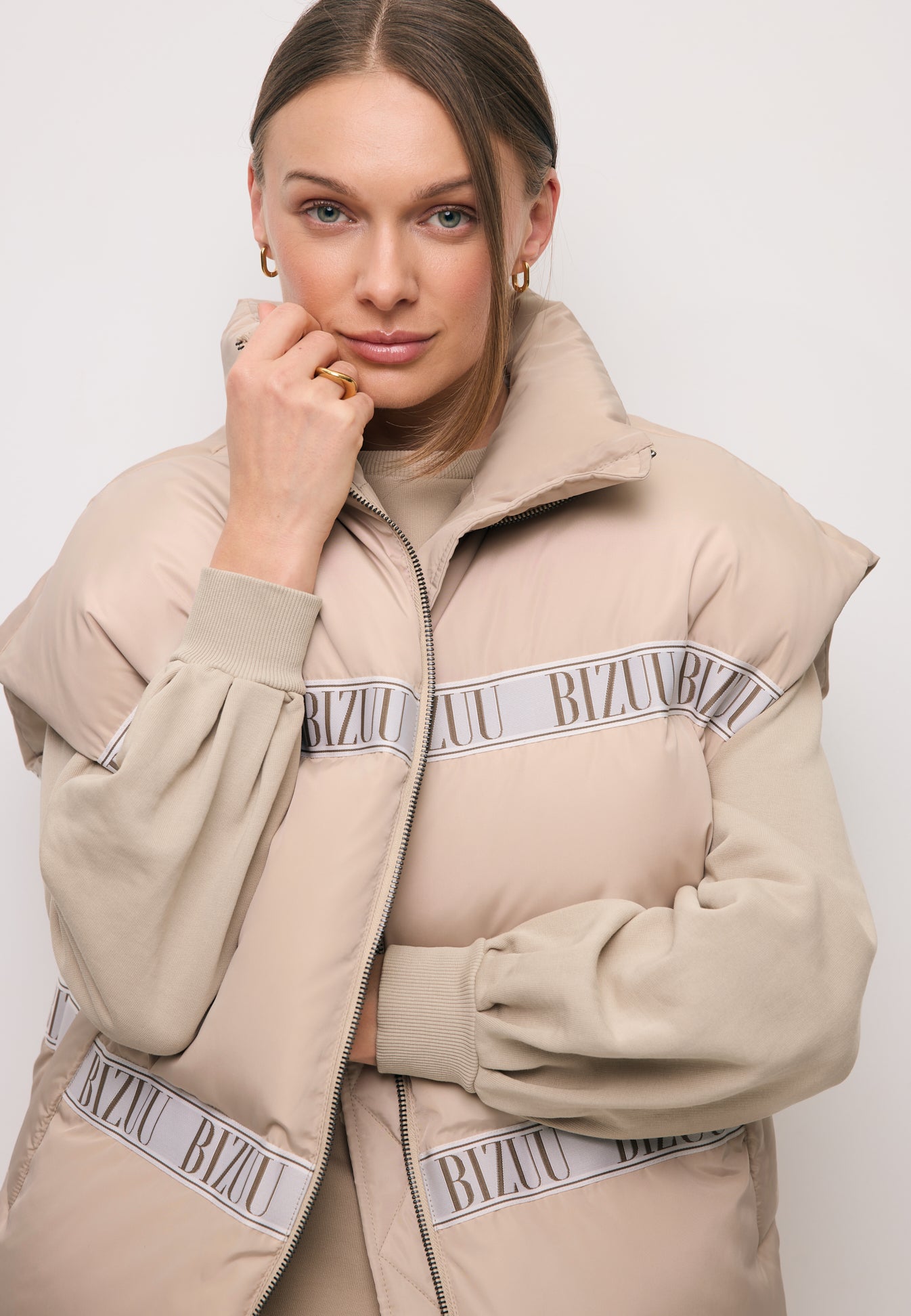 Zbliżenie na modelkę z ręką przy twarzy obraną w beżową kamizelkę puchową z logowaną taśmą AYASE.