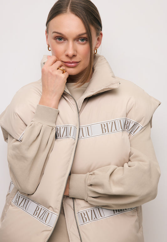 Zbliżenie na modelkę z ręką przy twarzy obraną w beżową kamizelkę puchową z logowaną taśmą AYASE.