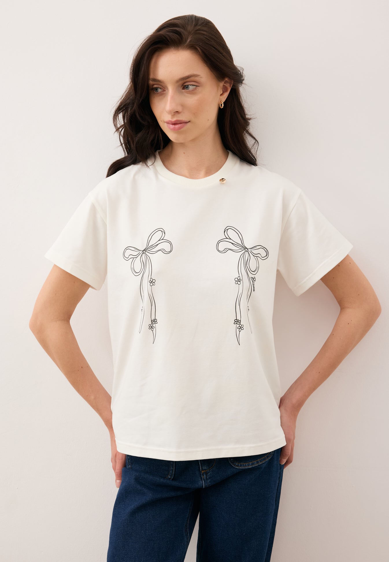 Modelka oparta o ścianę z rękoma ugiętymi na biodrach, ubrana w kremowy T-shirt z rysunkiem kokardek BOW oraz spodnie LEYA.