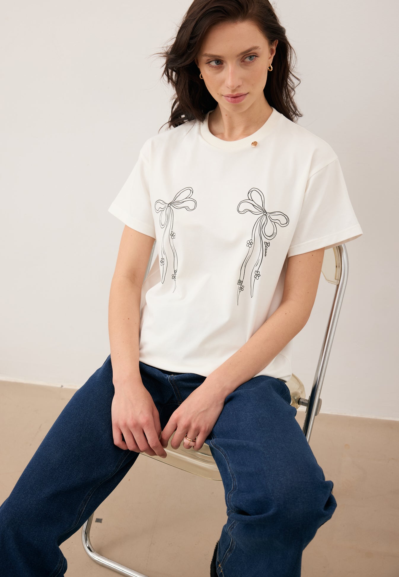 Modelka siedząca na krześle ubrana w kremowy T-shirt z kokardkami i logo BOW oraz granatowe jeansy LEYA.