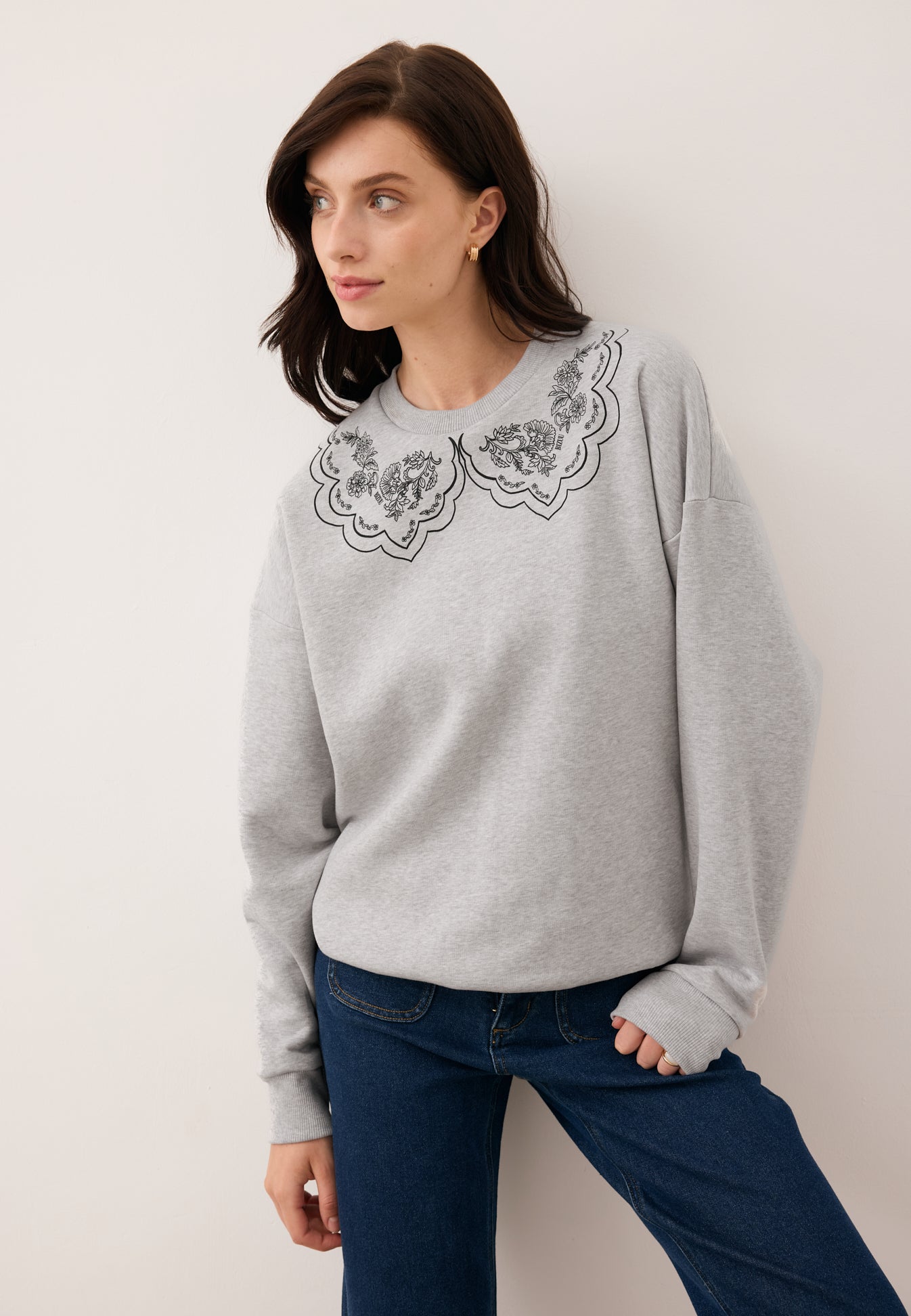 Modelka z opartą na nodze ręką ubrana w dzianinową bluzę COLLIER w kolorze szarym z rysunkiem kołnierza.