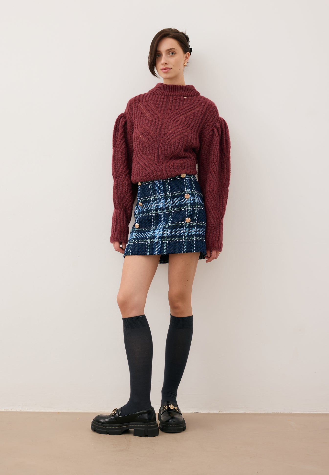Modelka oparta o ścianę, ubrana w mini spódniczkę w granatową kratę DISSA oraz bordowy sweter ROSSO.