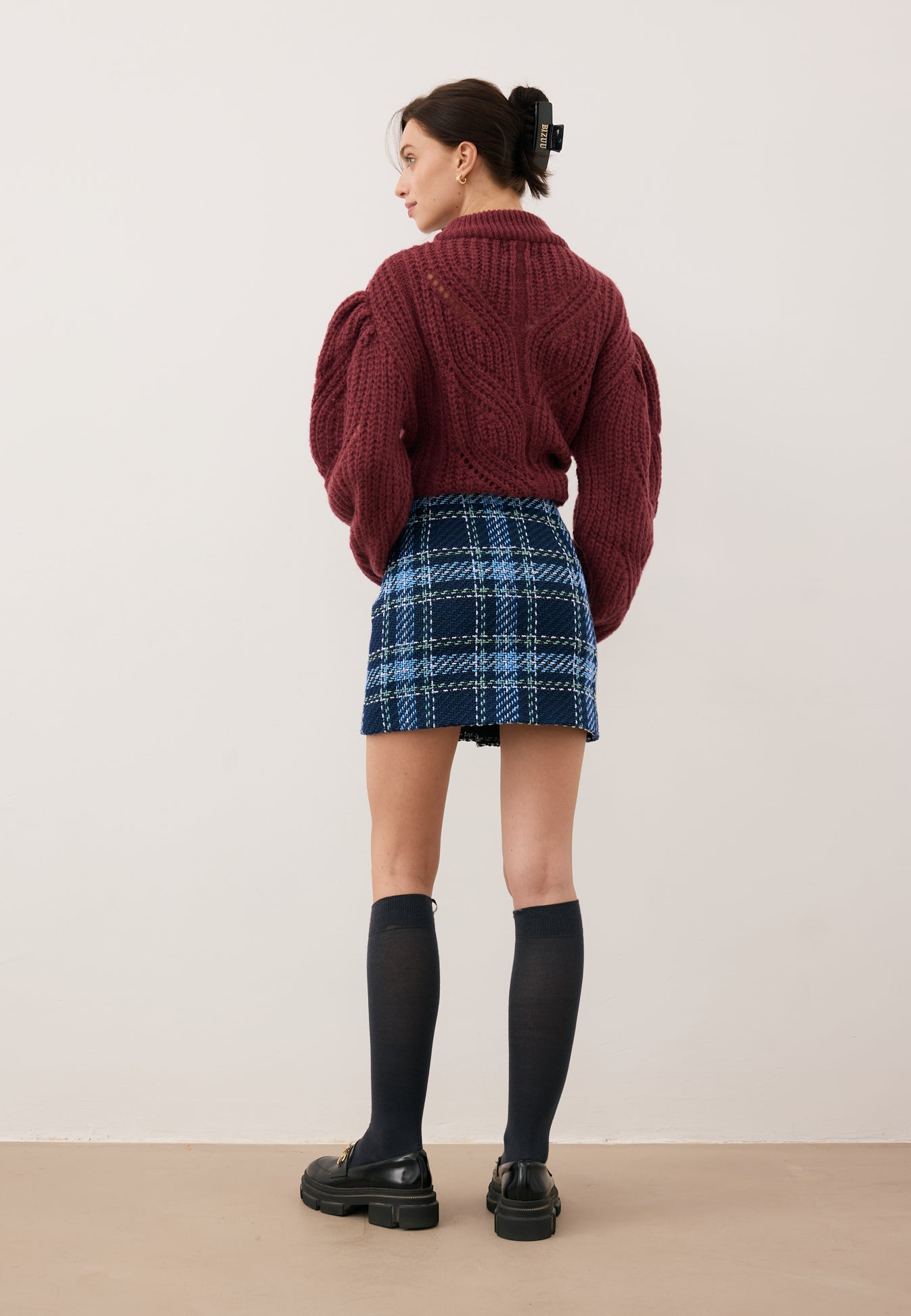 Modelka stojąca tyłem ubrana w bordowy sweter ROSSO, granatową mini spódniczkę DISSA, podkolanówki SCHOOLI i buty LESOTHO.