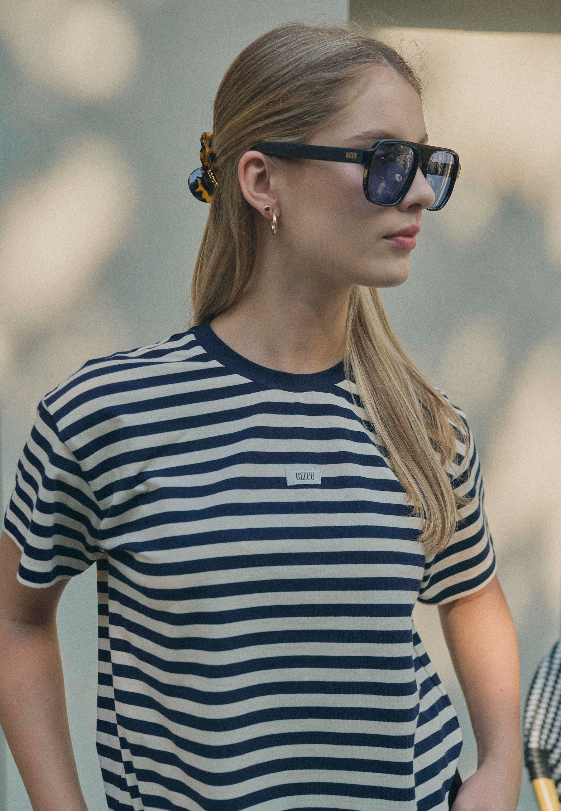 Zbliżenie na kobietę w czarnych okularach przeciwsłonecznych BIZUU z odbijającymi światło szkłami. Ma na sobie pasiasty t-shirt z haftem BIZUU oraz złote kolczyki. Włosy spięte dużą klamrą w odcieniach bursztynowego brązu.