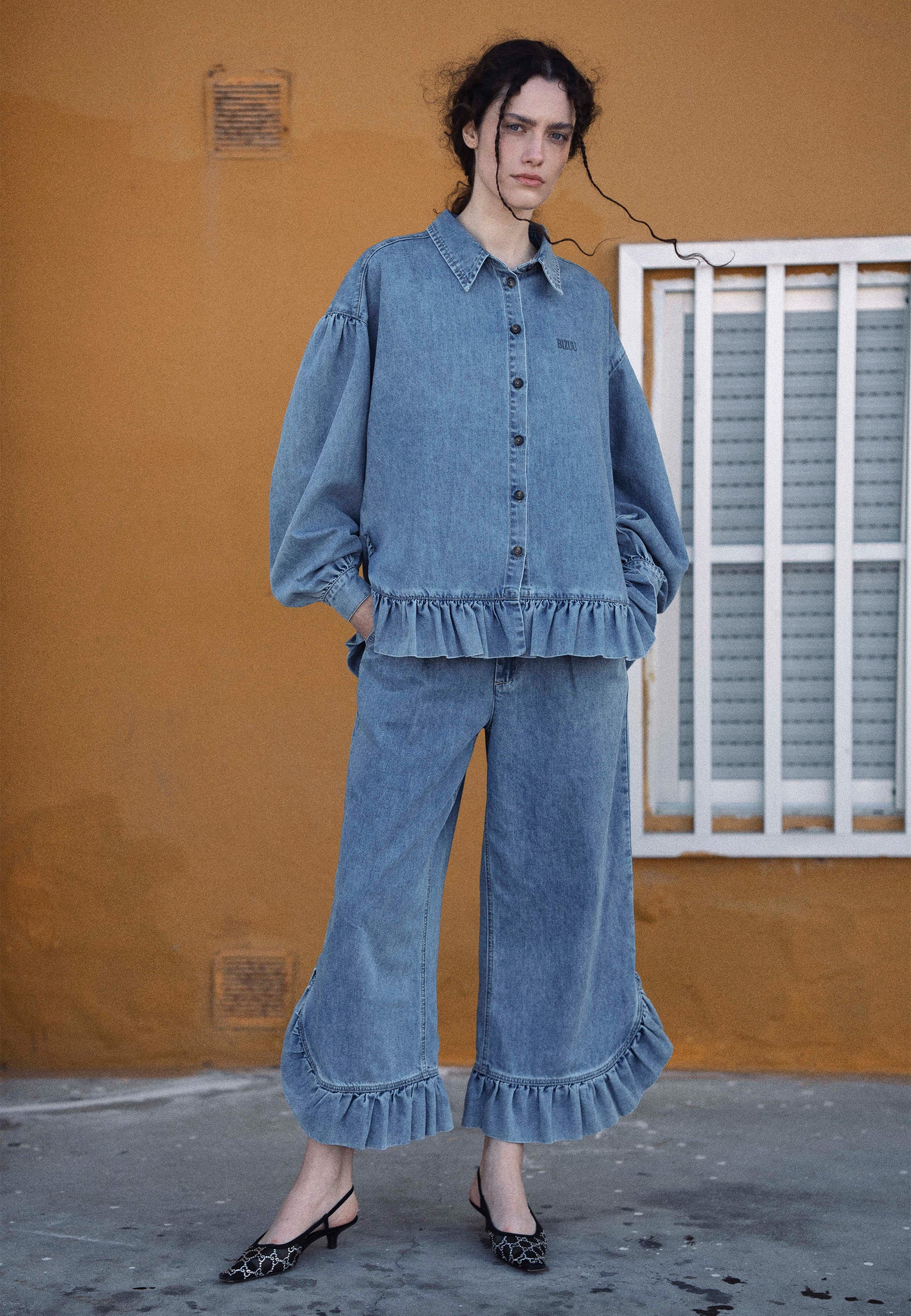 Modelka stojąca na ulicy ubrana w jeansowy zestaw z falbankami spodnie NELLA oraz koszulę FALBI.