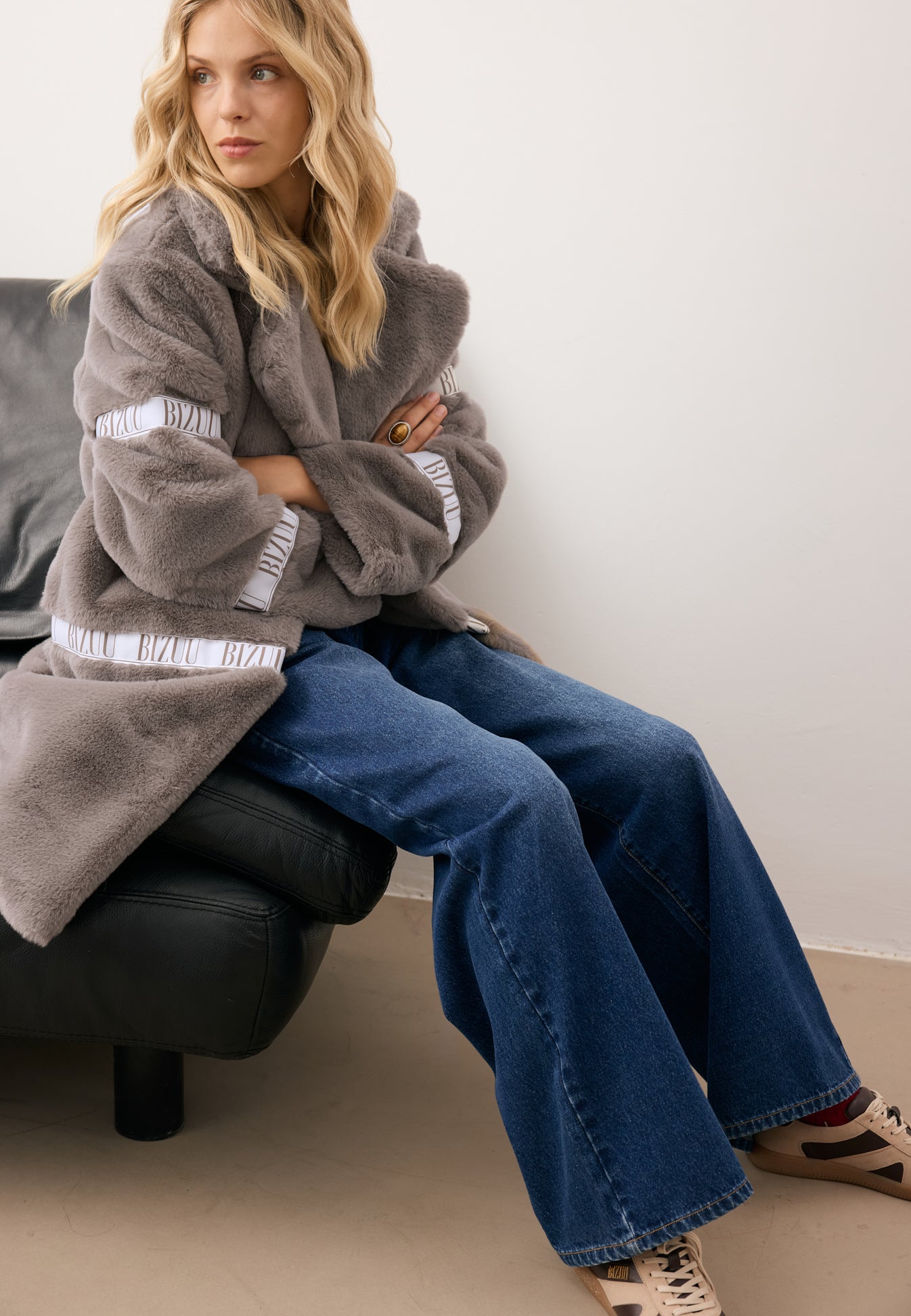 Modelka siedząca na kanapie z rękoma skrzyżowanymi na klatce piersiowej. Ubrana w płaszcz futrzany FLORENCE taupe i granatowe jeansy LEYA.