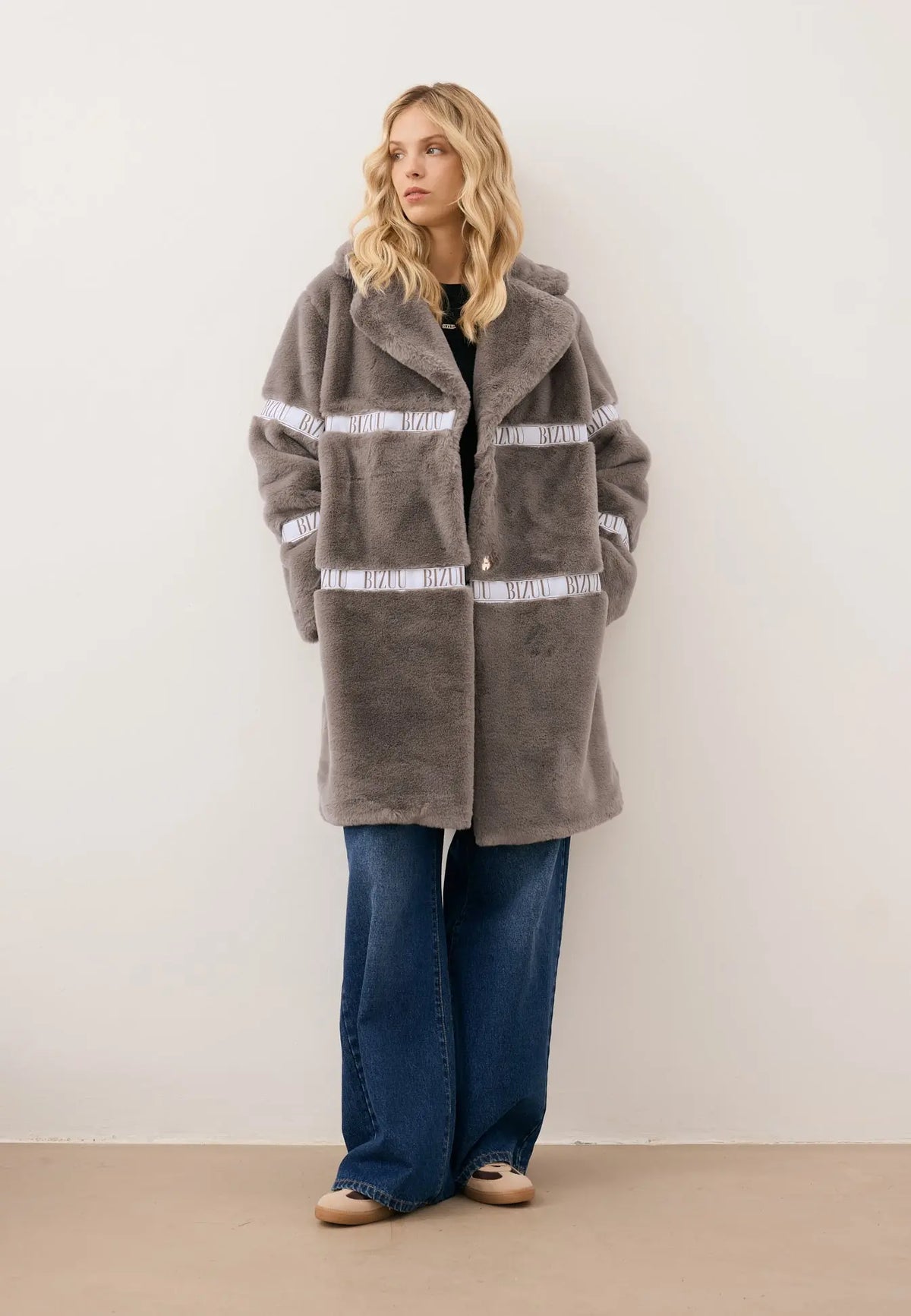 Modelka patrząca w bok, z rękoma w kieszeni futrzanego płaszcza FLORENCE z logo w kolorze taupe.