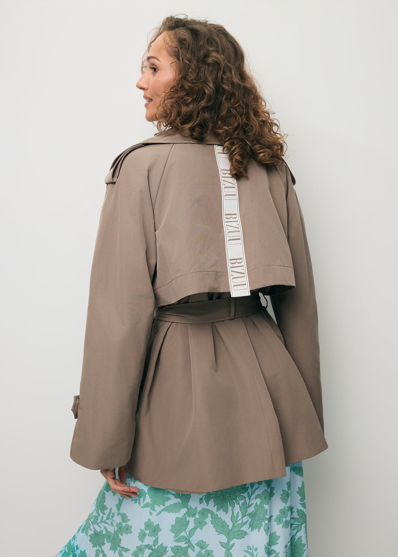 Modelka stojąca tyłem ubrana w krótki trencz z logowaną taśmą na plecach RACHY .