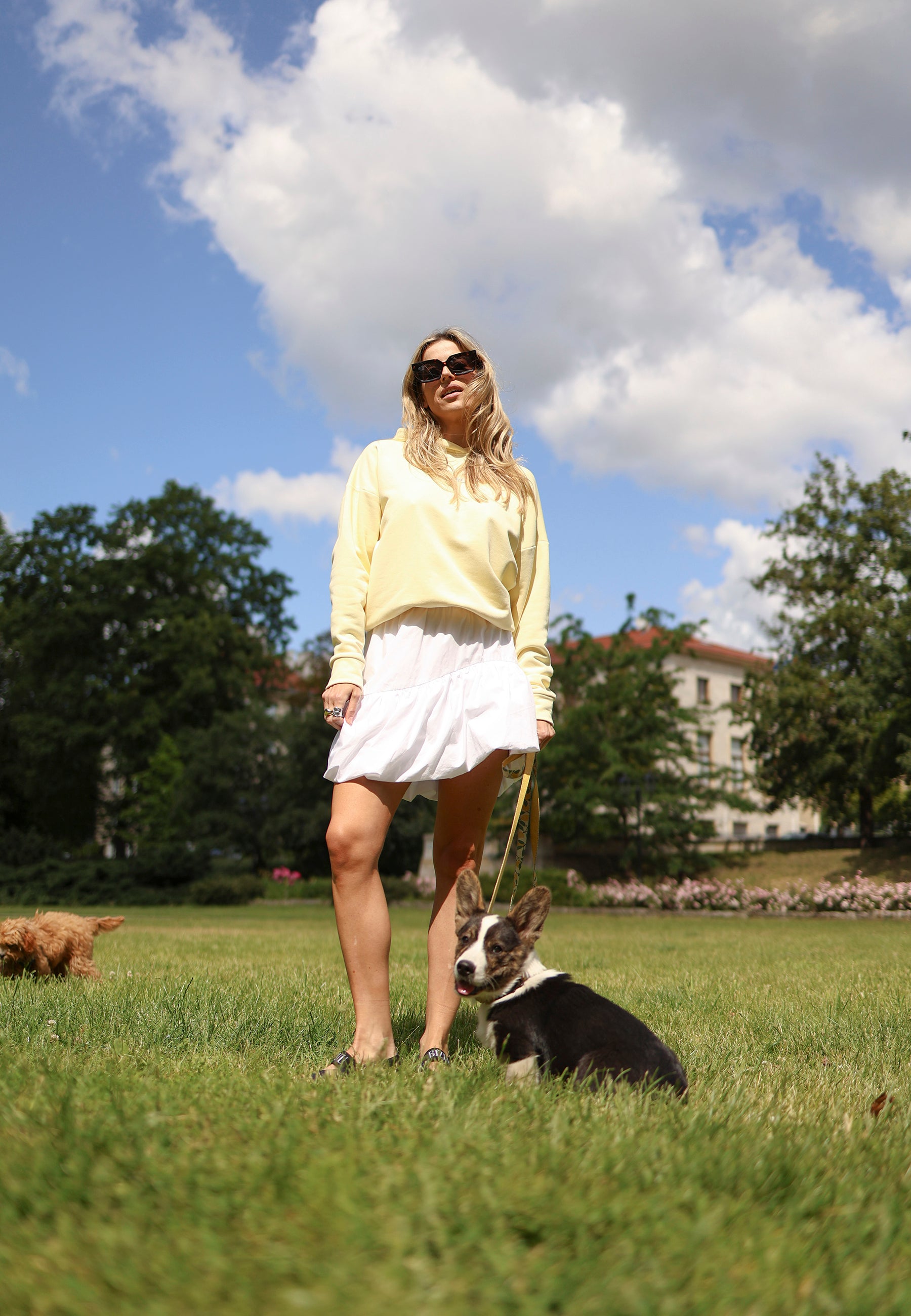 Modelka stojąca na trawie z psem ubrana w żółtą bluzę LINDA oraz białą mini spódniczkę LILIBET.