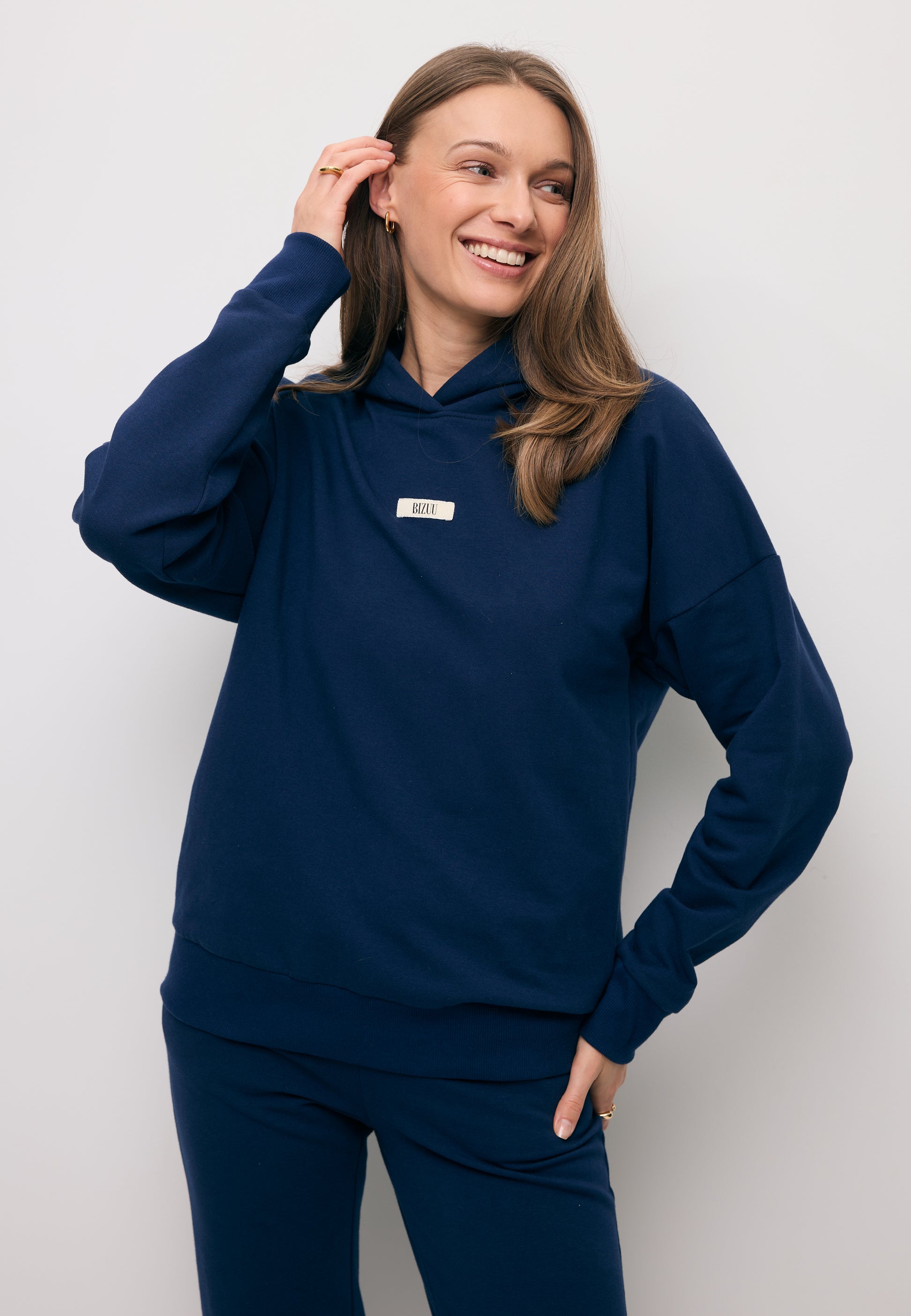 Zbliżenie na uśmiechniętą modelkę poprawiającą włosy ubraną w granatową bluzę z kapturem LINDI z logowaną metką na przodzie.