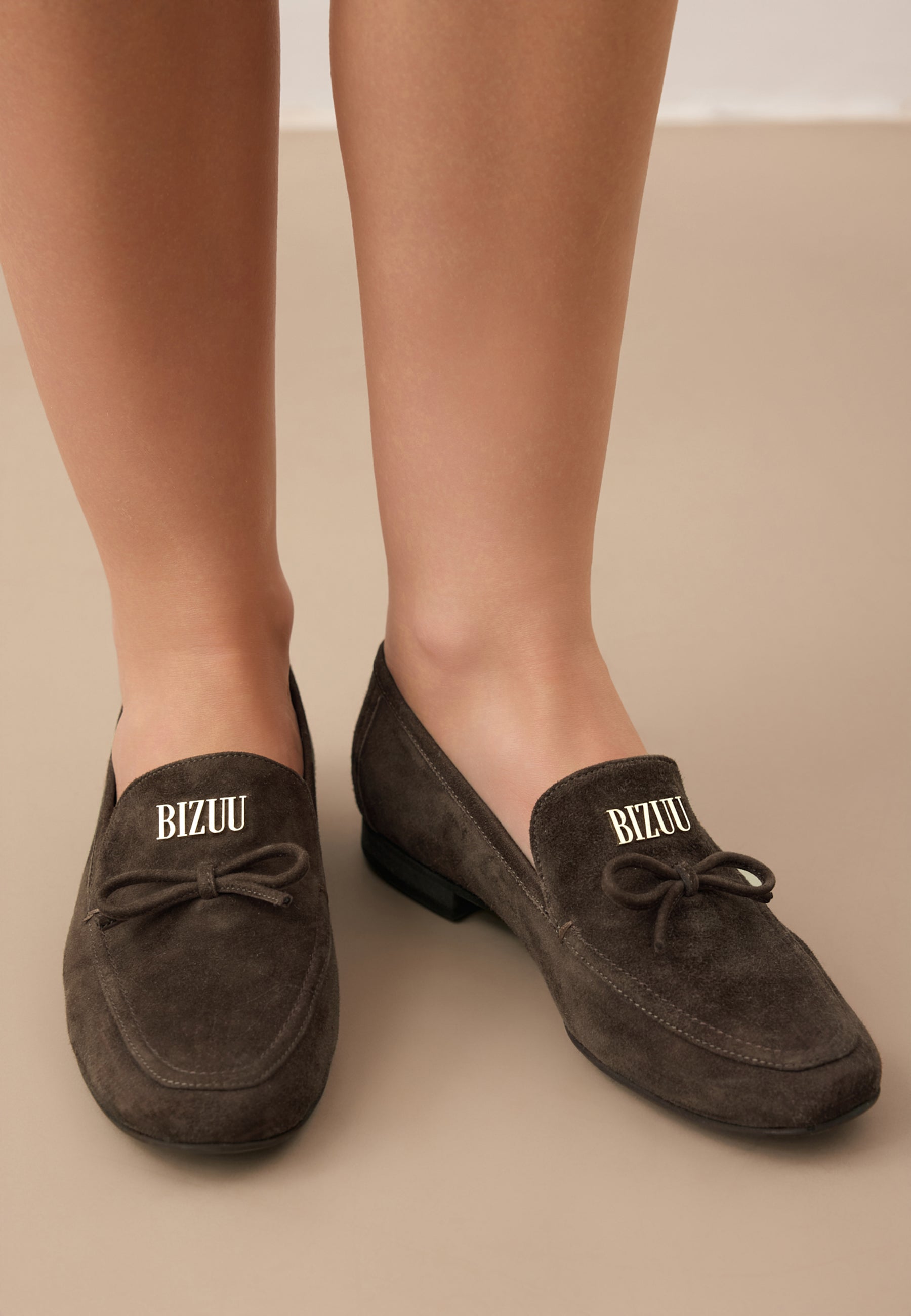 Zbliżenie na nogi modelki w brązowych, zamszowych mokasynach LISANA ze złotym logo na przodzie.