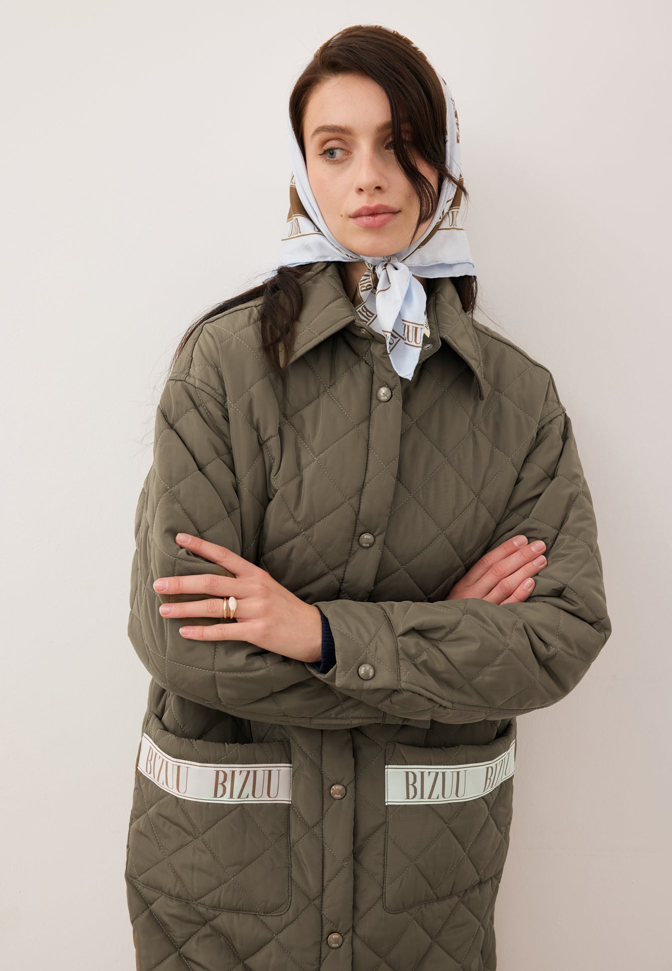Zbliżenie na modelkę w pikowanym płaszczu MAREVIA khaki z logowaną taśmą na kieszeniach, na głowie modelka przewiązaną ma niebieską apaszkę ARIA z jedwabiu.