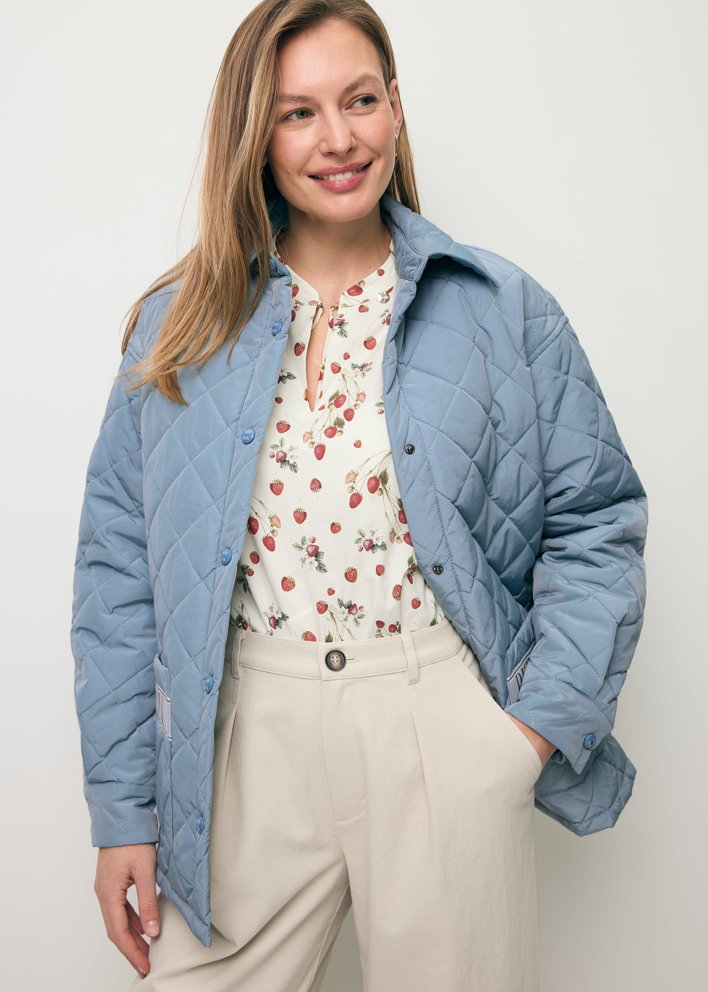 Uśmiechnięta modelka z ręką w kieszeni kremowych spodni NELLA, ubrana w błękitną kurtkę MARIN oraz bluzkę w poziomki ANDRASSA.