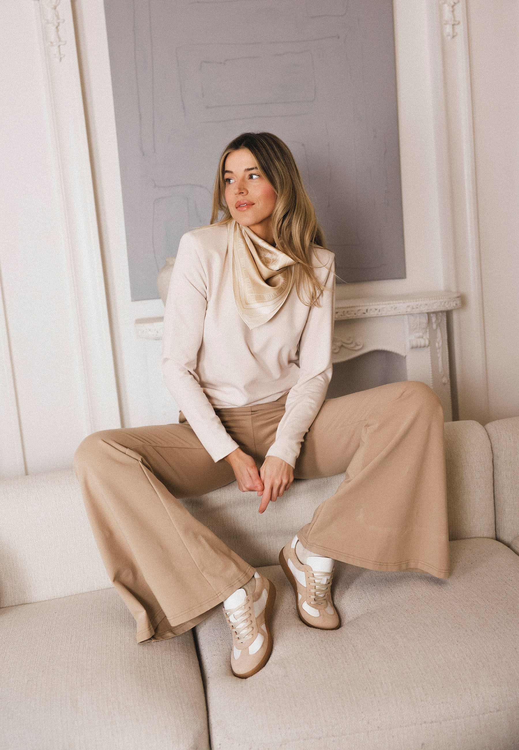 Modelka siedząca na kanapie ubrana w kremową bluzkę MELLISANDRA oraz beżowe spodnie dresowe TEIA.