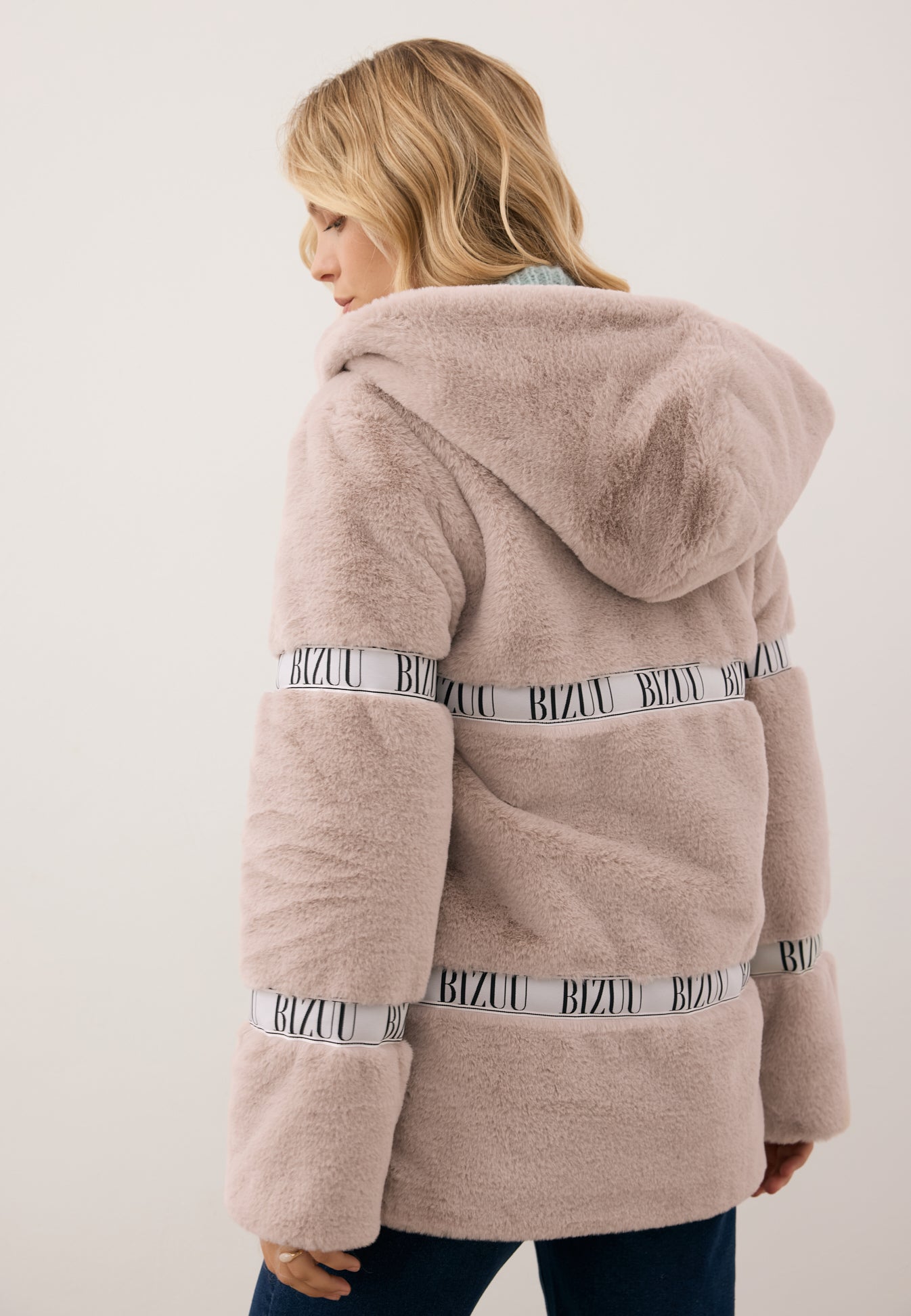 Modelka stojąca tyłem w beżowym płaszczyku z futra MILANO z kapturem oraz logowaną taśmą.