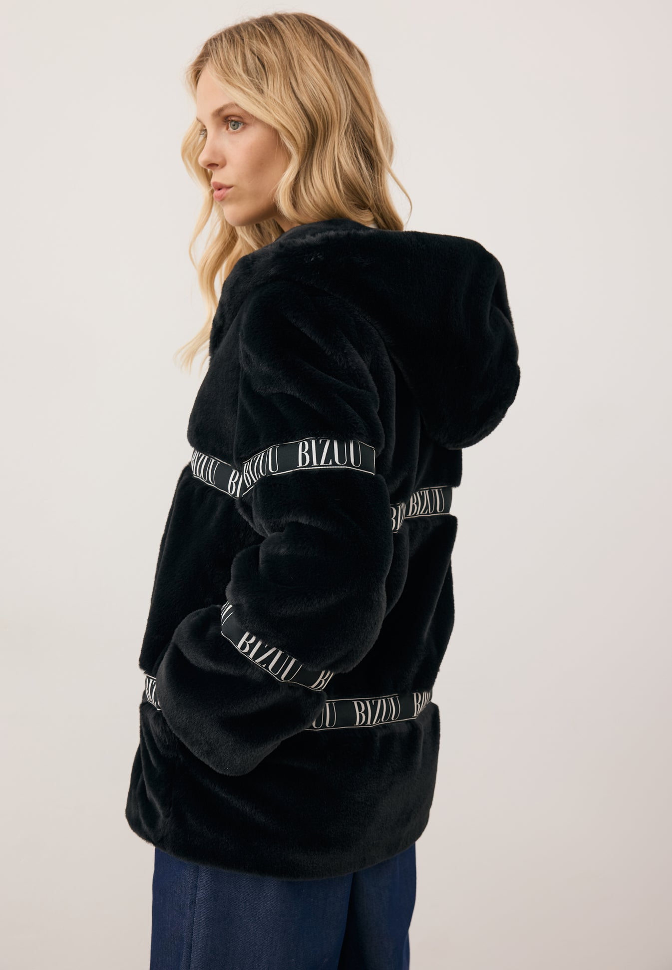 Modelka stojąca bokiem z jedną ręką w kieszeni czarnego futrzanego płaszcza z logowanymi taśmami i kapturem MILANO.