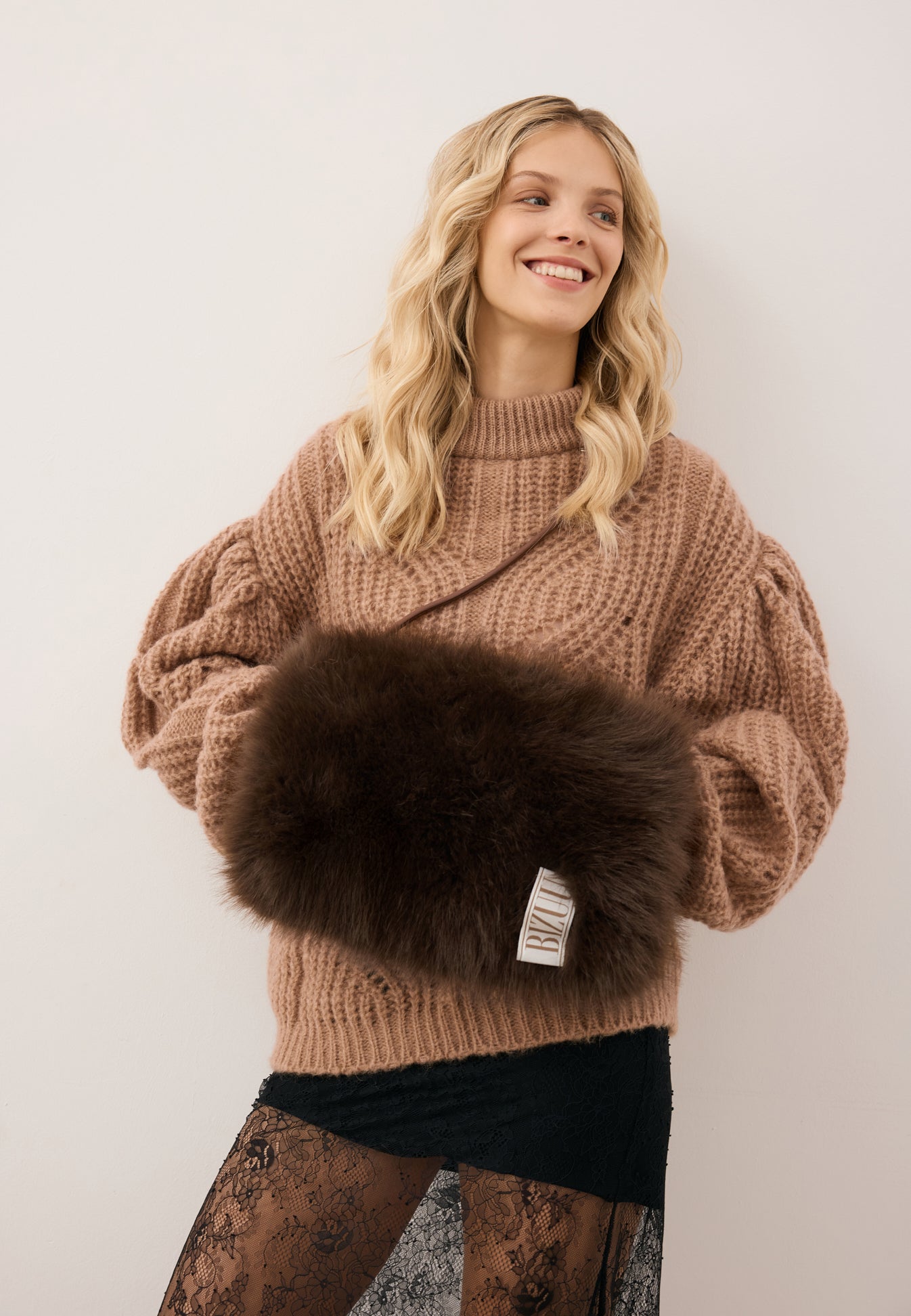 Uśmiechnięta modelka z rękoma w futrzanej muffce MUFFIE w kolorze brązowym.