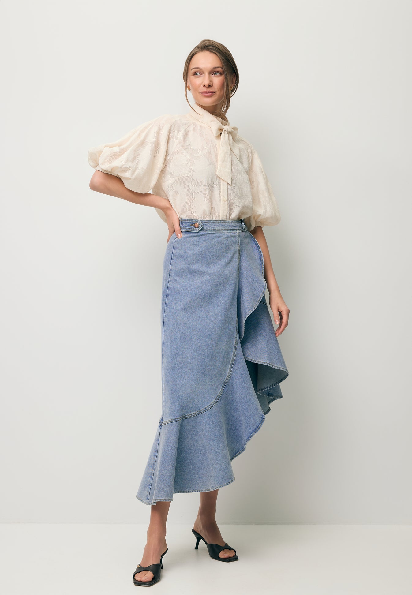 Modelka stojąca po skosie ubrana w jeansową spódniczkę z ozdobną falbaną NIIA oraz koszulą w kwiatowy wzór AZALEA w kolorze kremowym.