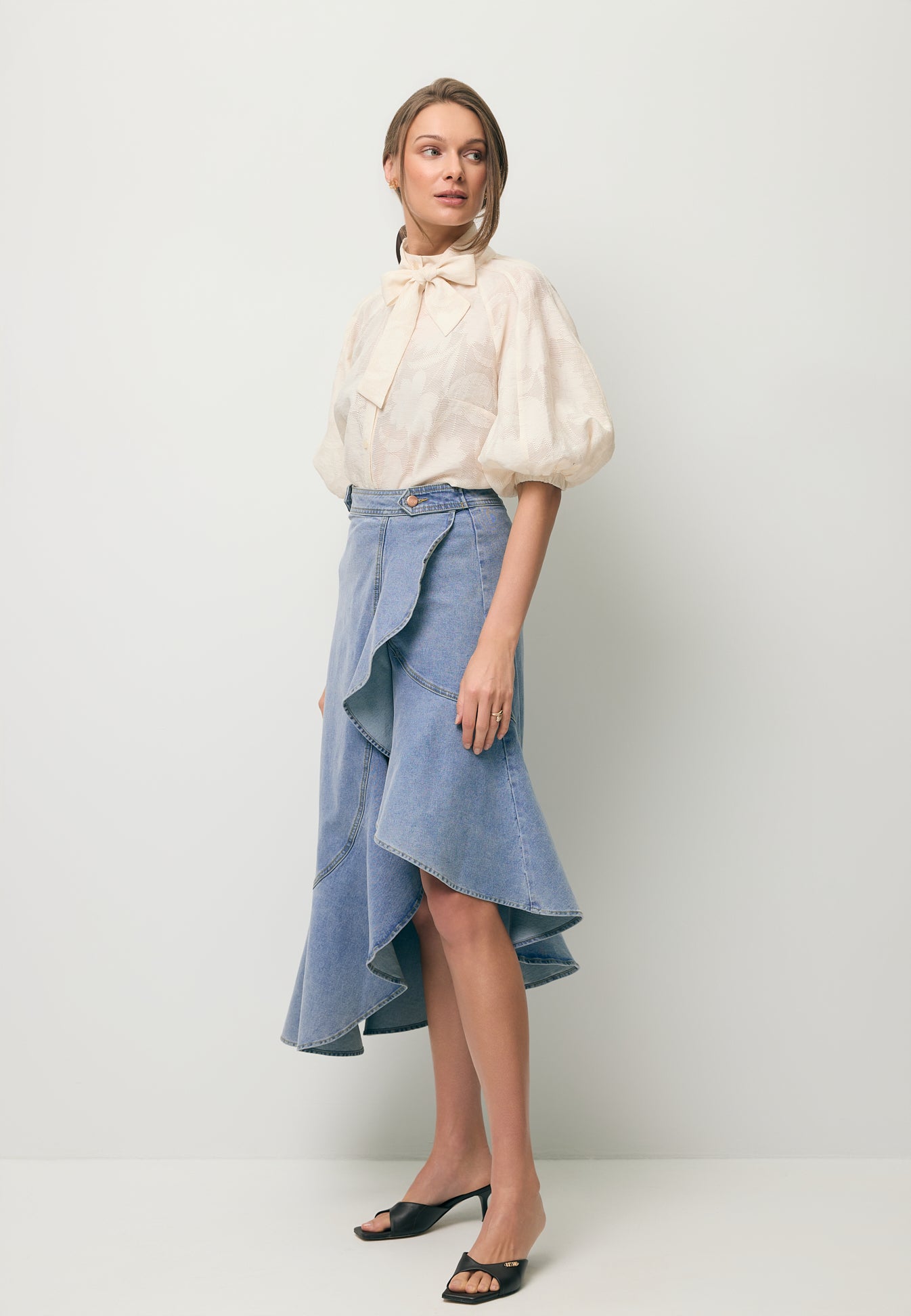 Modelka stojąca po skosie prezentująca asymetrię w spódniczce jeansowej NIIA z ozdobną falbaną na dole. Do tego ubraną ma koszulę w kwiatowy wzór AZALEA.