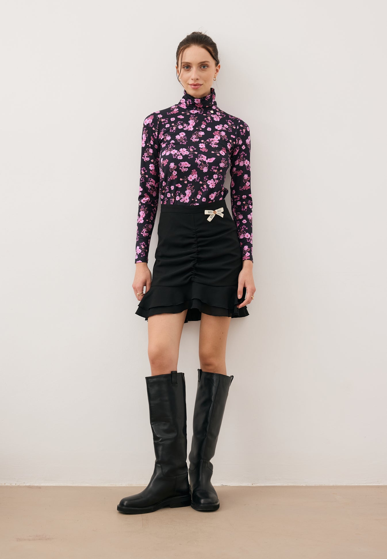 Modelka stojąca na wprost w dopasowanym golfie w kwiatowy print OWAKA, czarnej mini spódniczce ROSALIAN oraz kozakach CALYX.