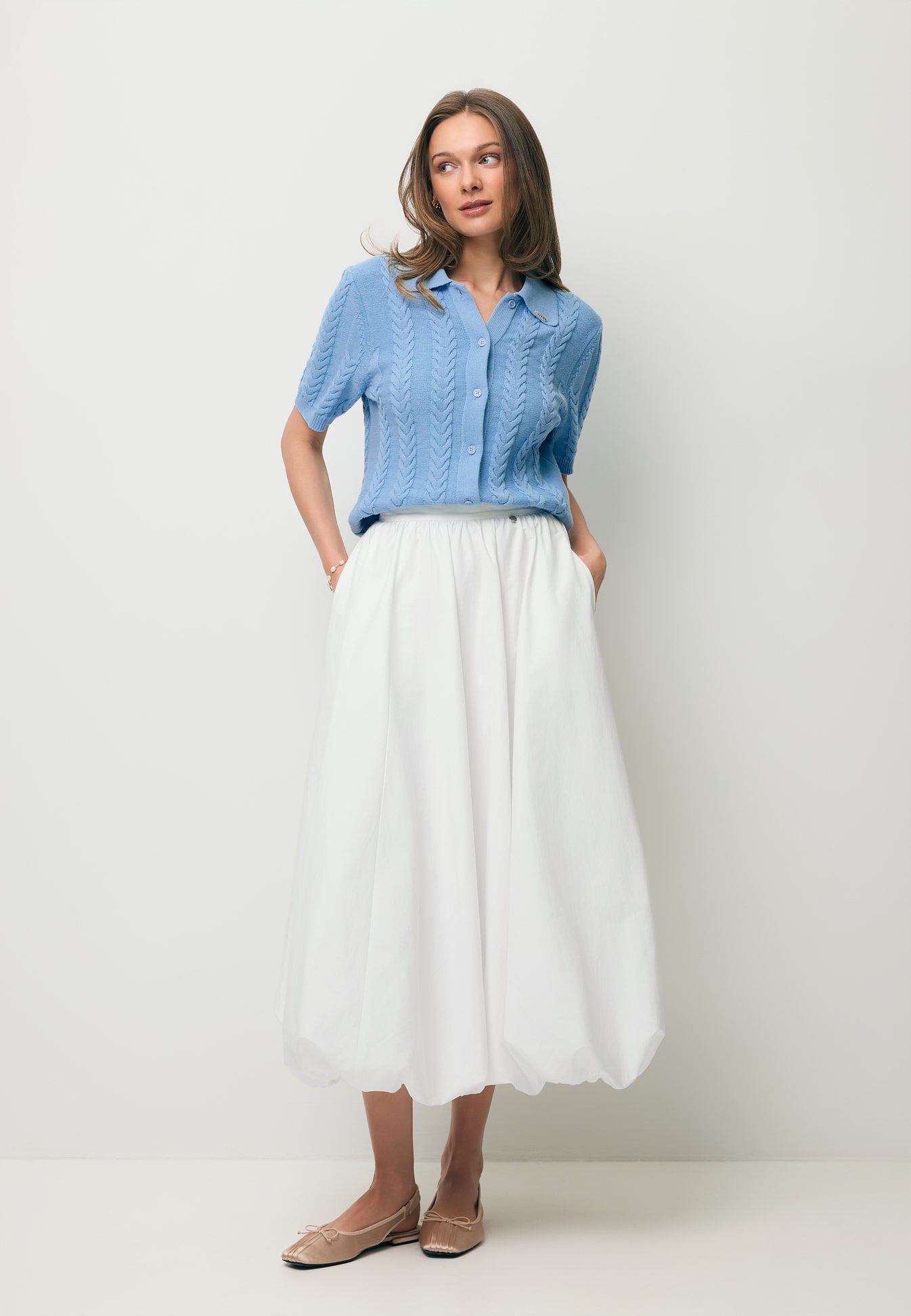 Modelka z rękoma w kieszeni kremowej spódniczki NUBE, ubrana w błękitny sweterek z kołnierzykiem PATRI.