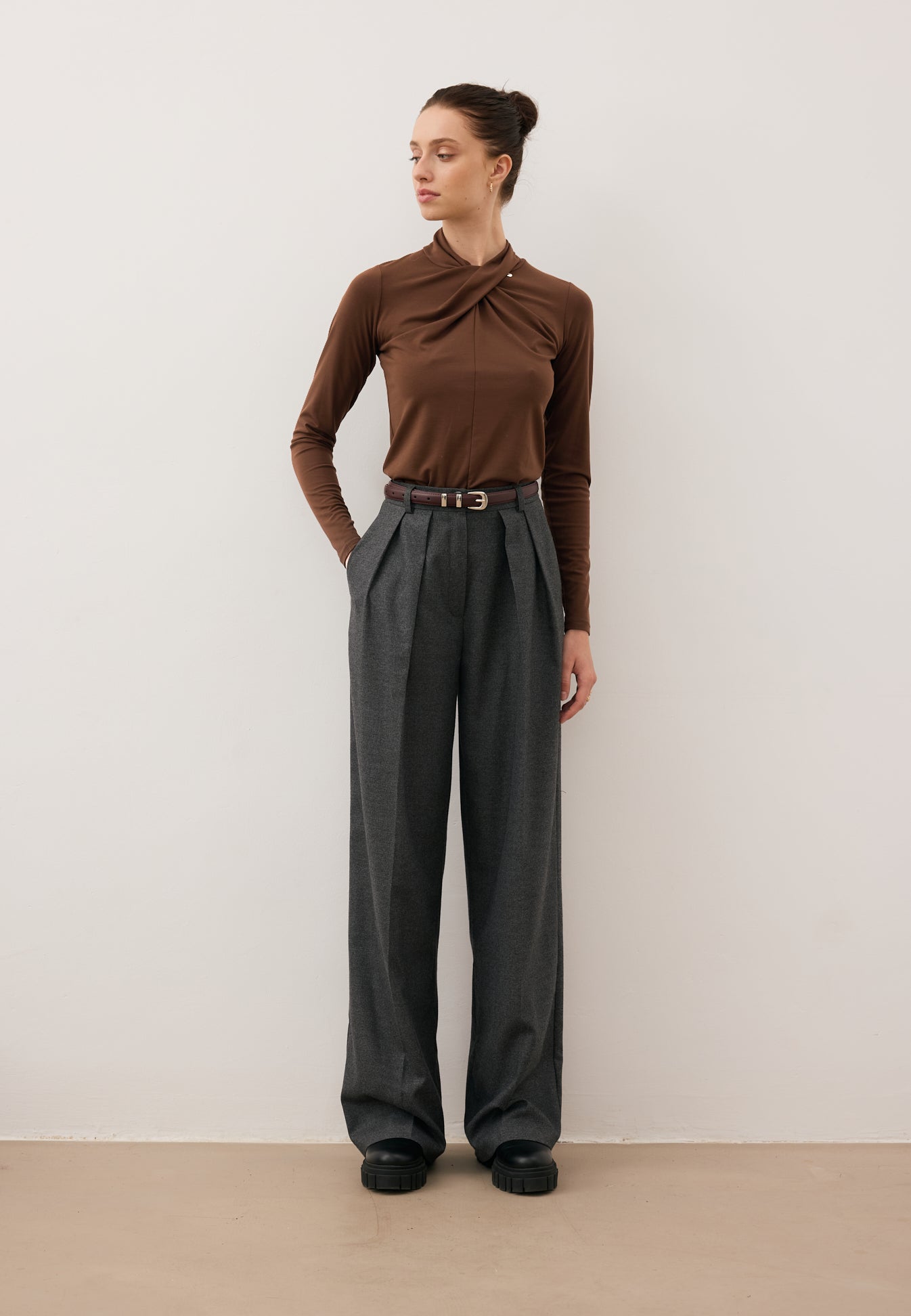 Modelka z jedną ręką w kieszeni spodni BANOS spogląda w bok. Ubrana w brązowy longsleeve PINOWA z zabudowanym dekoltem.
