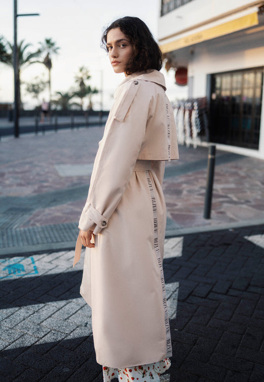 Modelka stojąca bokiem na przejściu dla pieszych ubrana w beżowy trencz z logowaną taśmą na plecach RACHELLA.