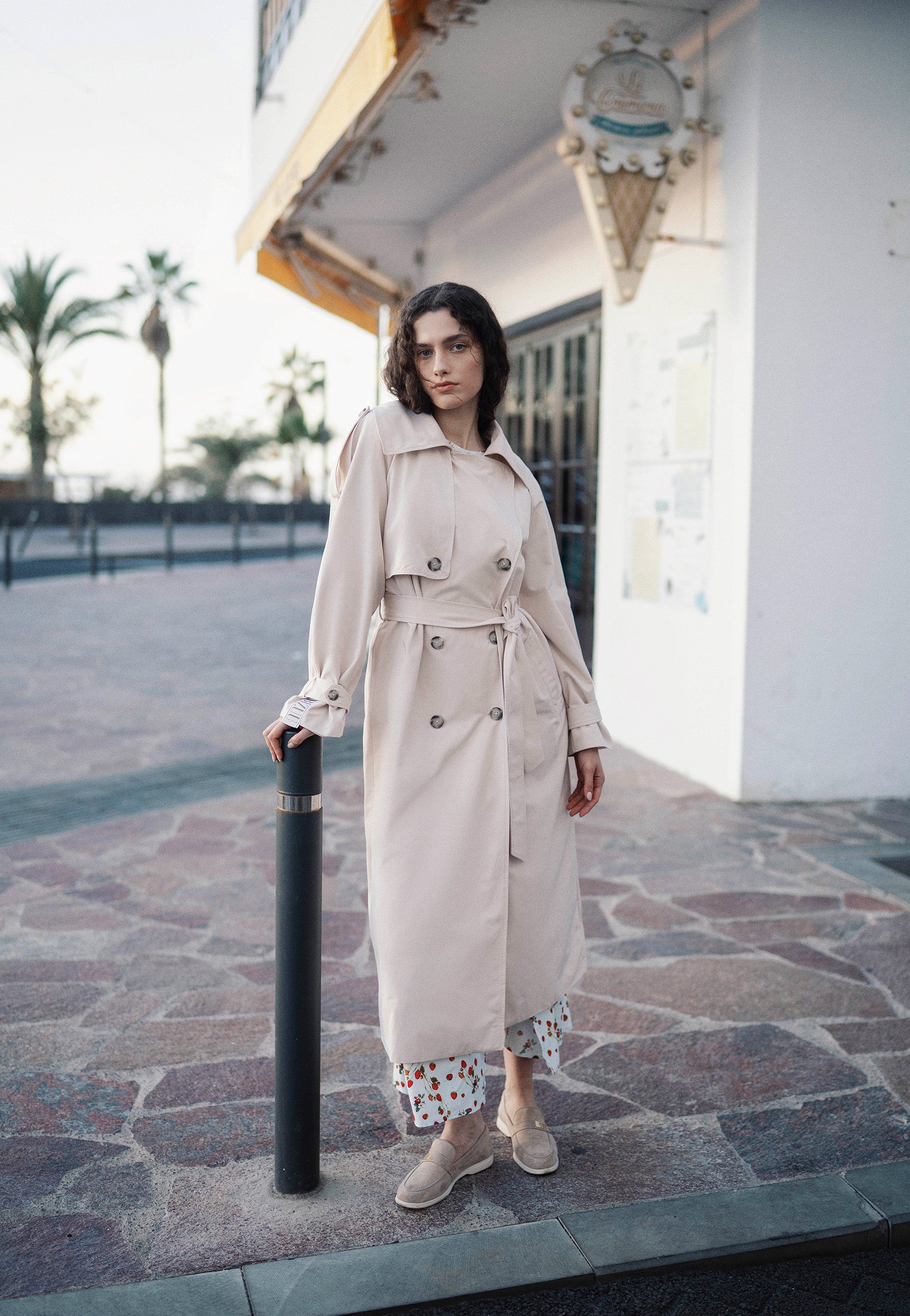 Modelka oparta o słupek ubrana w beżowy trencz RACHELLA z logowaną taśmą na plecach.