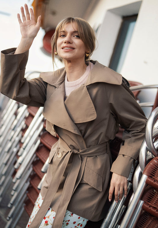 Modelka z podniesioną ręką ubrana w krótki płaszcz w kolorze taupe RACHY z taśmą z logo na plecach.