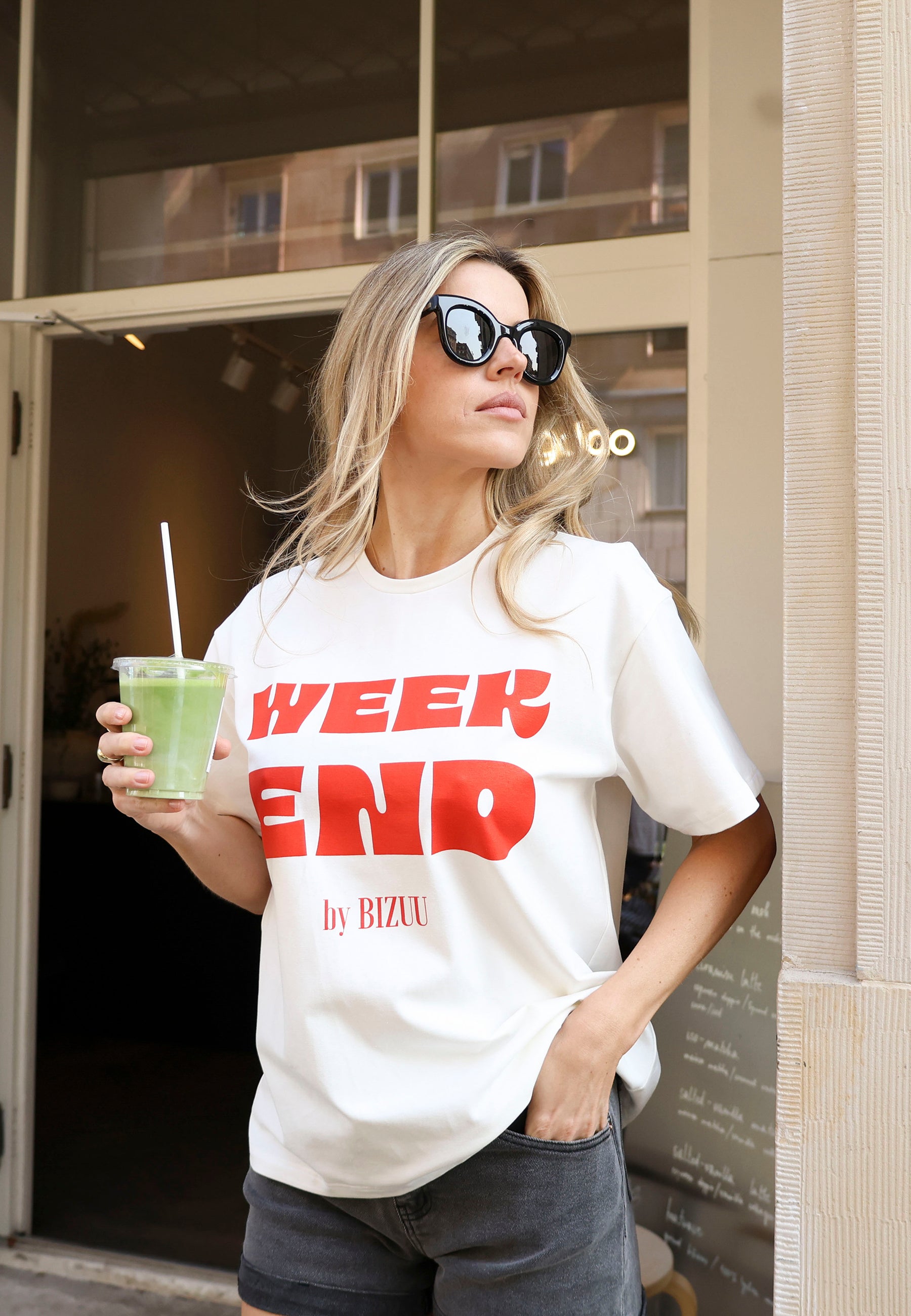 Zbliżenie na modelkę z jedną ręką w kieszeni jeansowych szortów MENORCA trzymająca w drugiej ręce napój. Ubrana w biały T-shirt z czerwonym napisem WEEKEND by Bizuu RALZA.