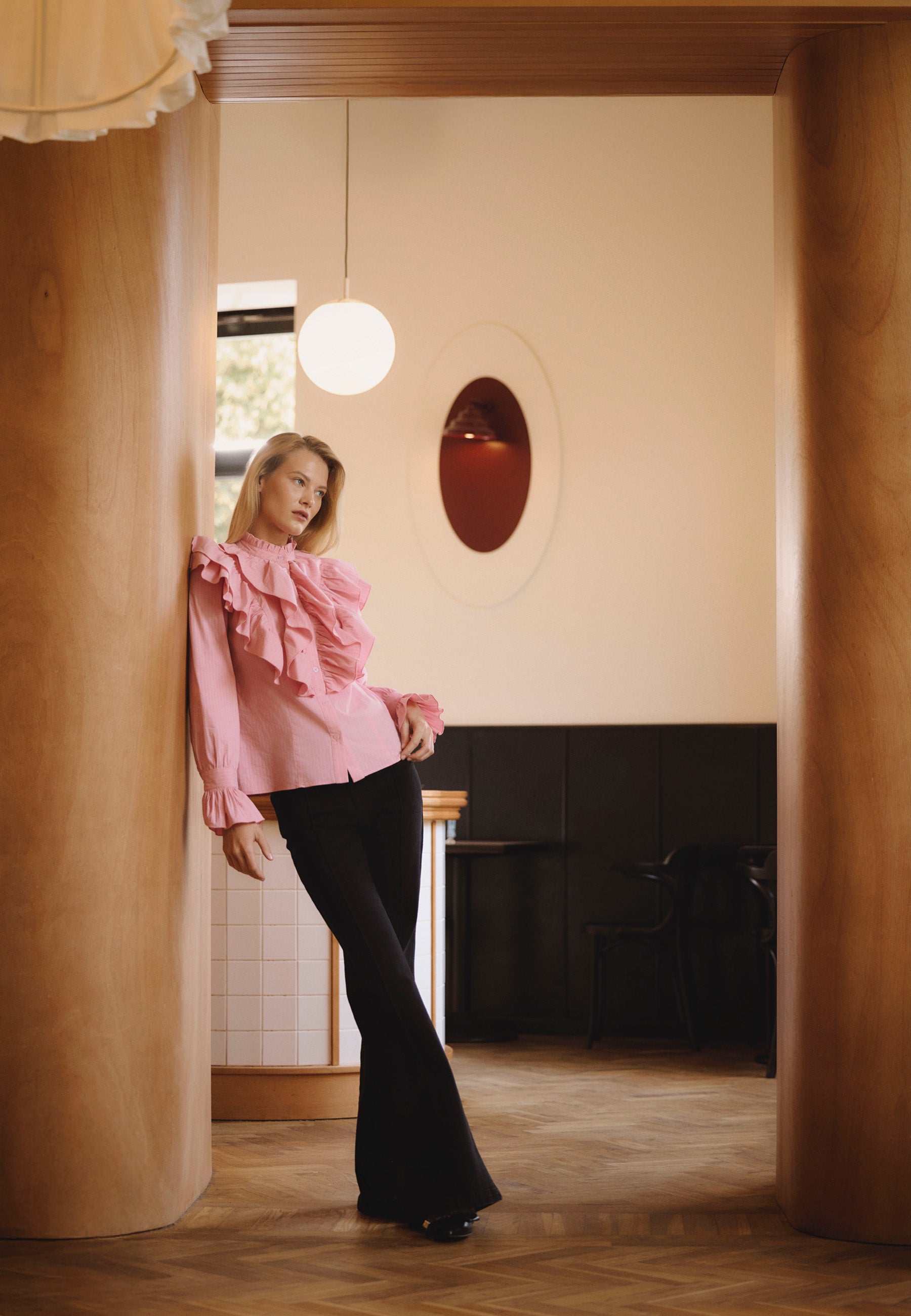 Modelka w kawiarni opiera się o drewnianą ścianę, stoi ze skrzyżowanymi nogami. Ubrana w różową koszulę z falbanami SANNI i czarne jeansy typu flare BELLINA.