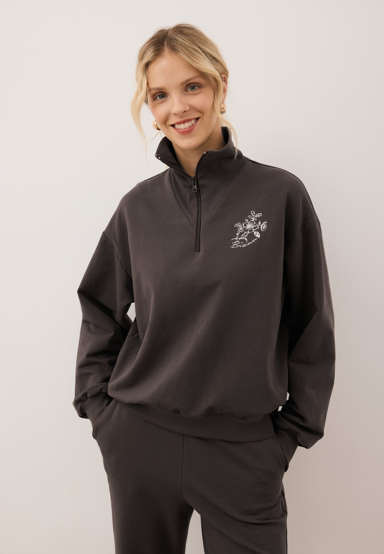 Uśmiechnięta modelka patrząca w kamerę, z rękoma w kieszeni spodni ZEIA. Zbliżenie na brązową bluzę TANSY z aplikacją z logo na przodzie.