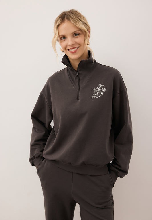 Uśmiechnięta modelka patrząca w kamerę, z rękoma w kieszeni spodni ZEIA. Zbliżenie na brązową bluzę TANSY z aplikacją z logo na przodzie.