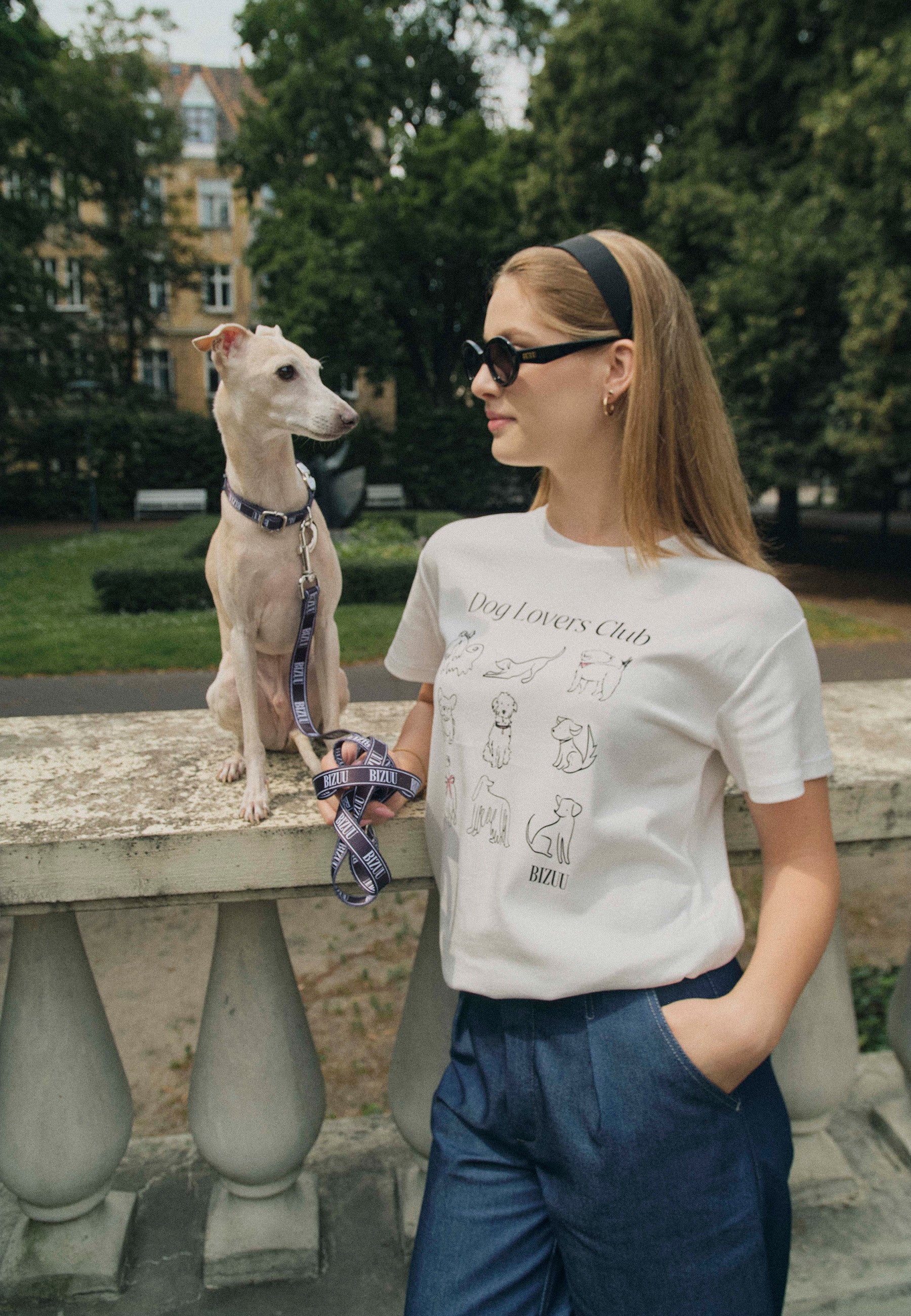 Modelka oparta o murek, na którym siedzi pies. Ubrana w biały T-shirt z rysunkami psów i napisem Dog Lovers Club. W ręce trzyma smycz w logo Bizuu.