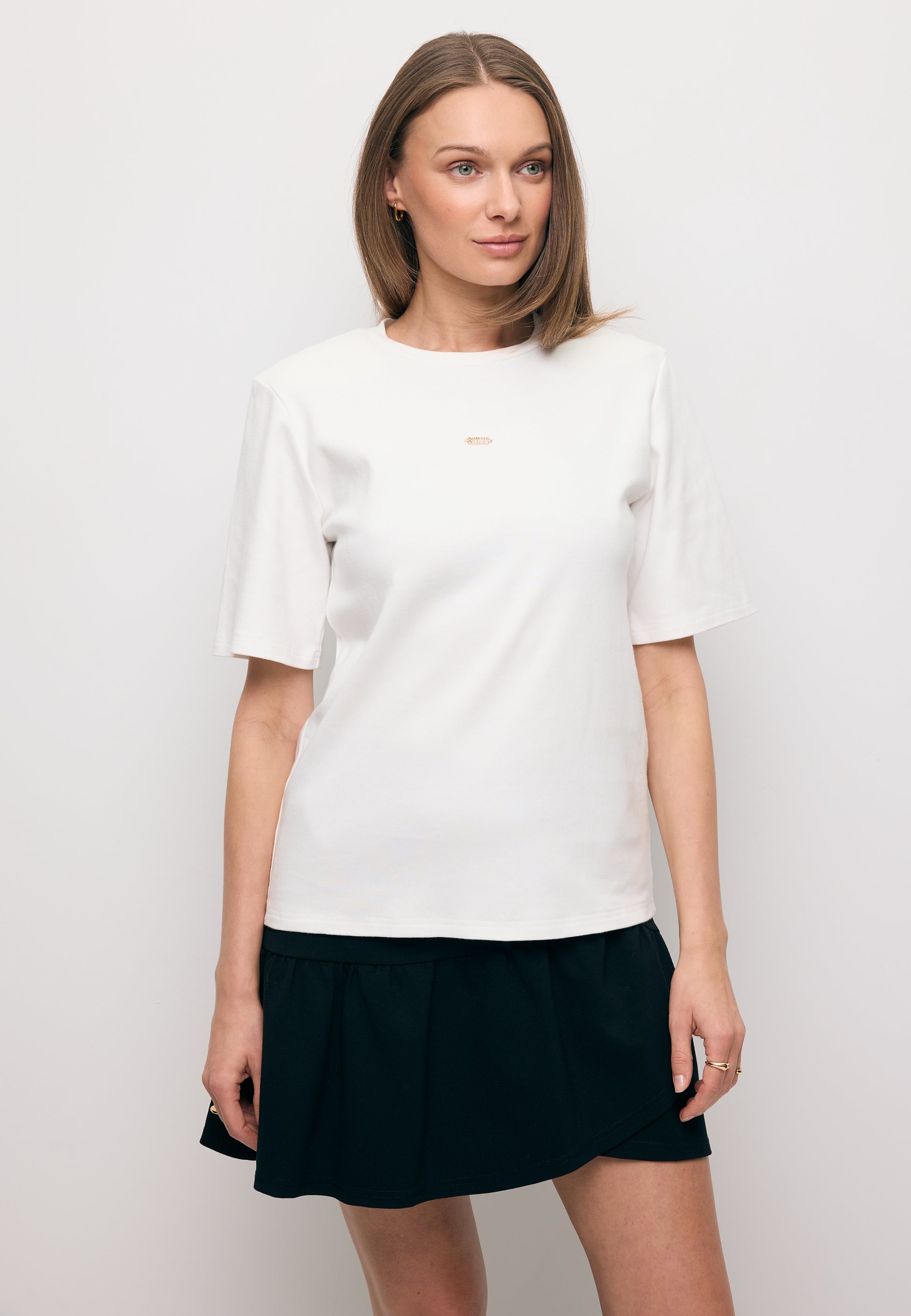 Modelka ubrana w białą koszulkę z ozdobnymi poduszkami MELISA oraz mini spódniczkę z falbankami TULIAN.