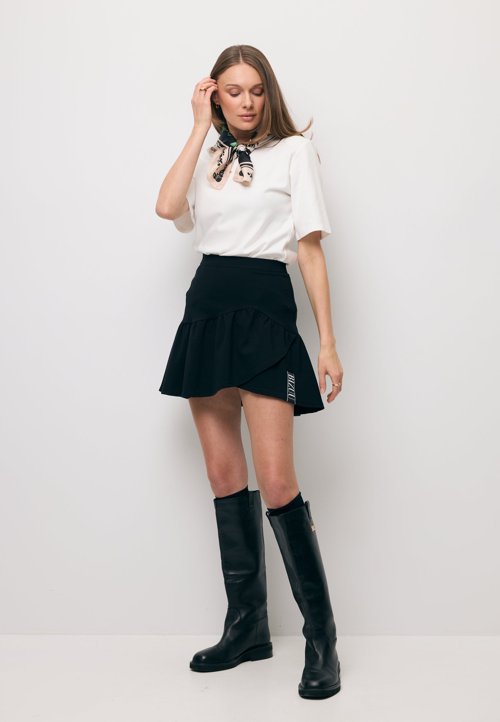 Modelka na tle białej ściany ubrana w T-shirt kremowy z ozdobnym logo i poduszkami MELISA oraz mini spódniczkę w kolorze czarnym TULIAN.