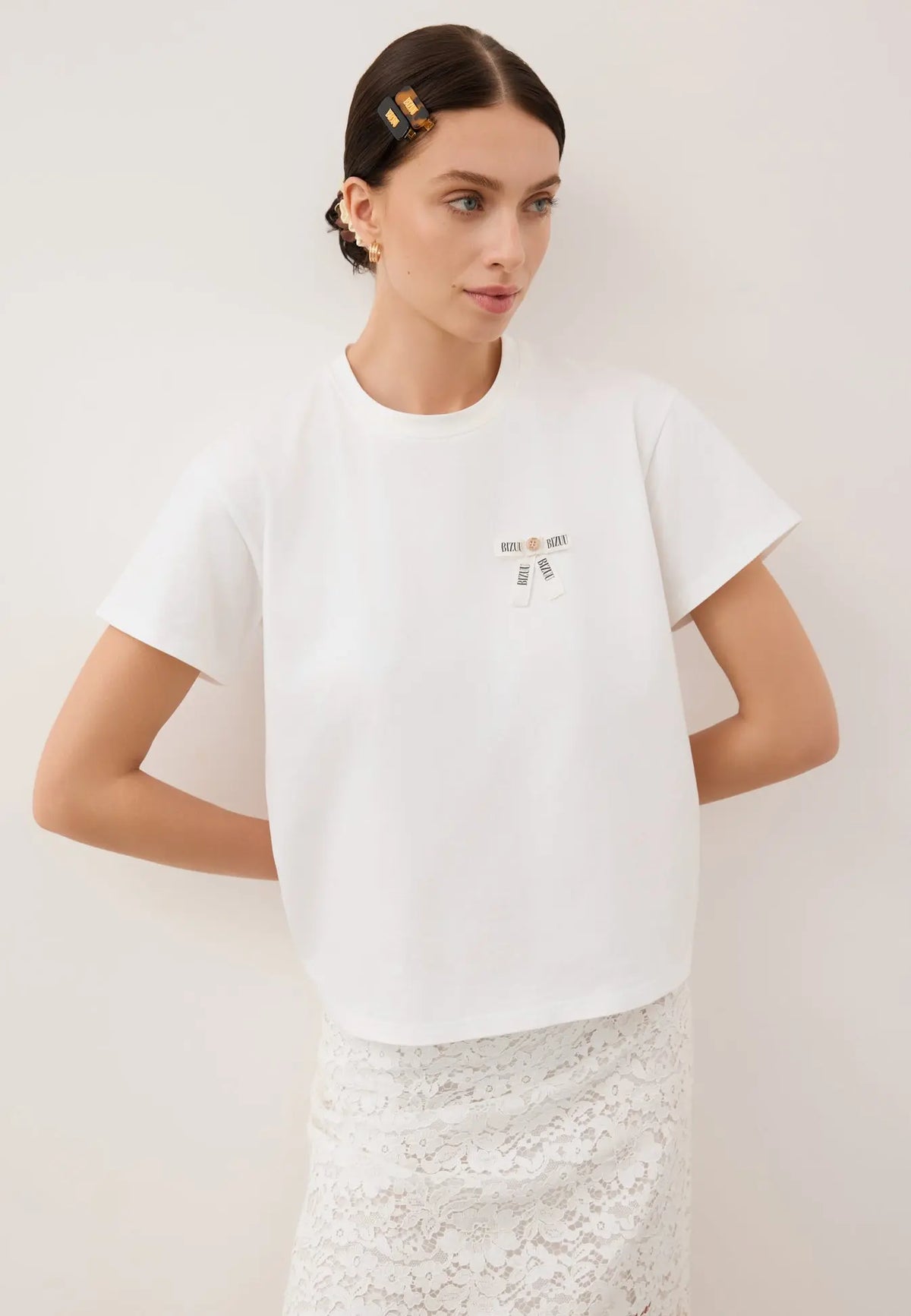 Modelka oparta o białą ścianę z rękoma z tyłu. Ubrana w biały T-shirt z logowaną kokardką TISHALLE i białą, koronkową spódnicę MILKY.