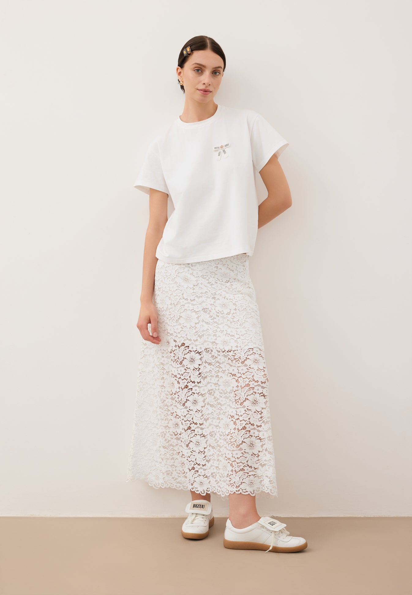 Modelka oparta o białą ścianę, ubrana w biały T-shirt z logowaną kokardką TISHALLE i białą, koronkową spódniczkę MILKY.