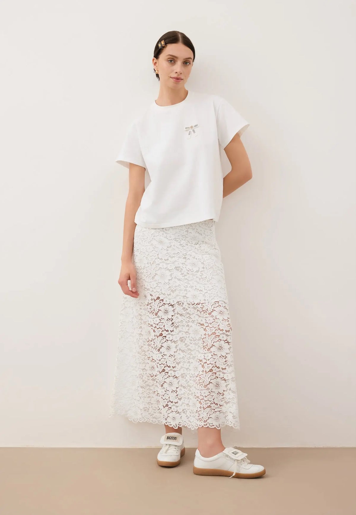Modelka oparta o białą ścianę, ubrana w biały T-shirt z logowaną kokardką TISHALLE i białą, koronkową spódniczkę MILKY.