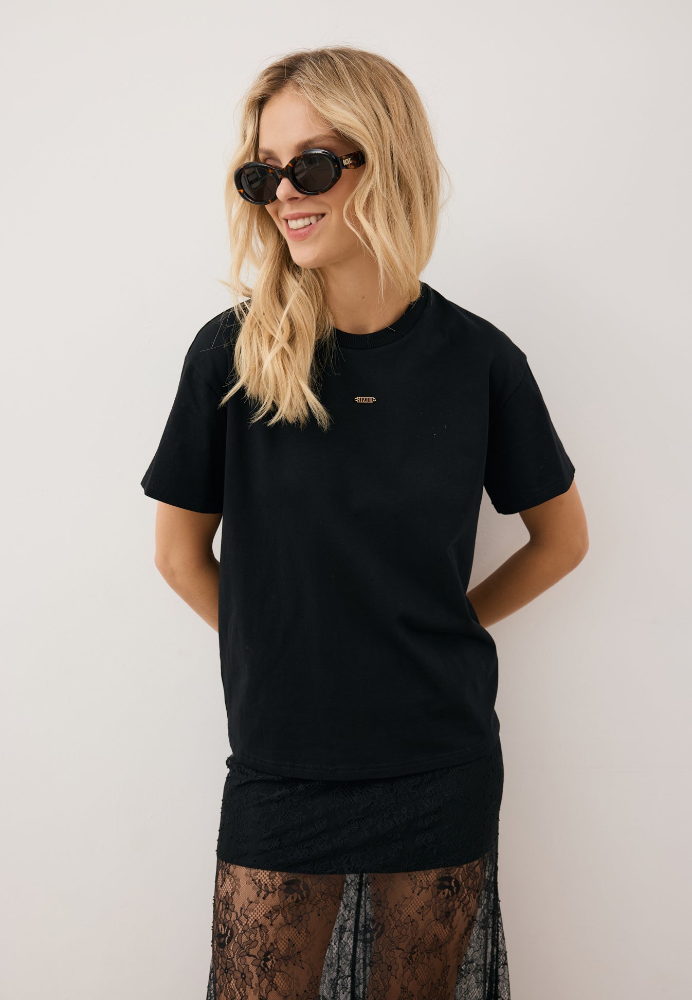 Modelka oparta o ścianę ubrana w czarny T-shirt TISHAL, czarną, koronkową spódnicę FLACE oraz okulary przeciwsłoneczne.