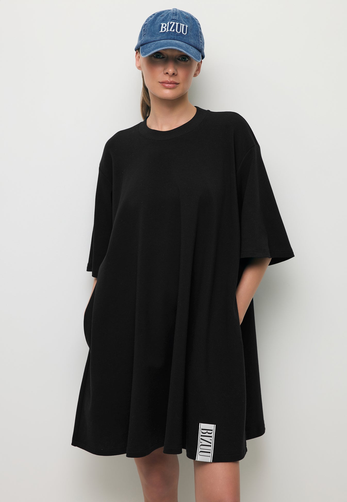 Zbliżenie na modelkę w mini dzianinowej sukience TRESS ozdobną logowaną metką oraz niebieską czapką z daszkiem ZANE.