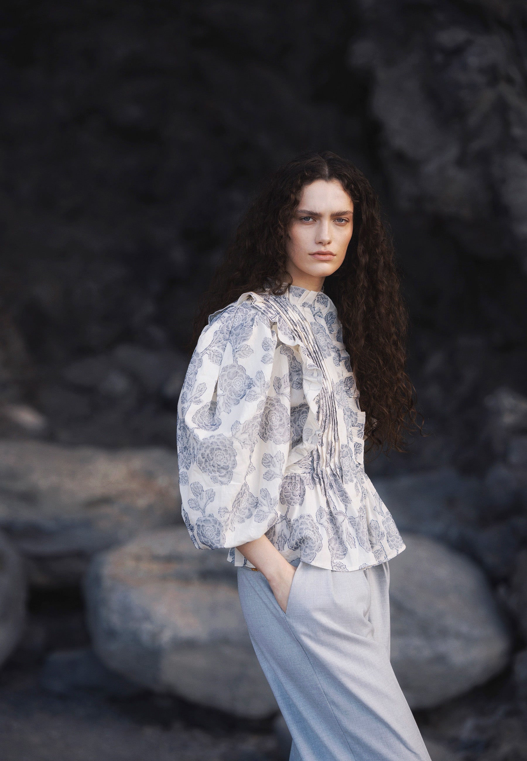 Modelka na tle skał ubrana w wzorzystą bluzkę z ozdobnymi falbanami ULLA oraz szare spodnie z szerokimi nogawkami BANOS.