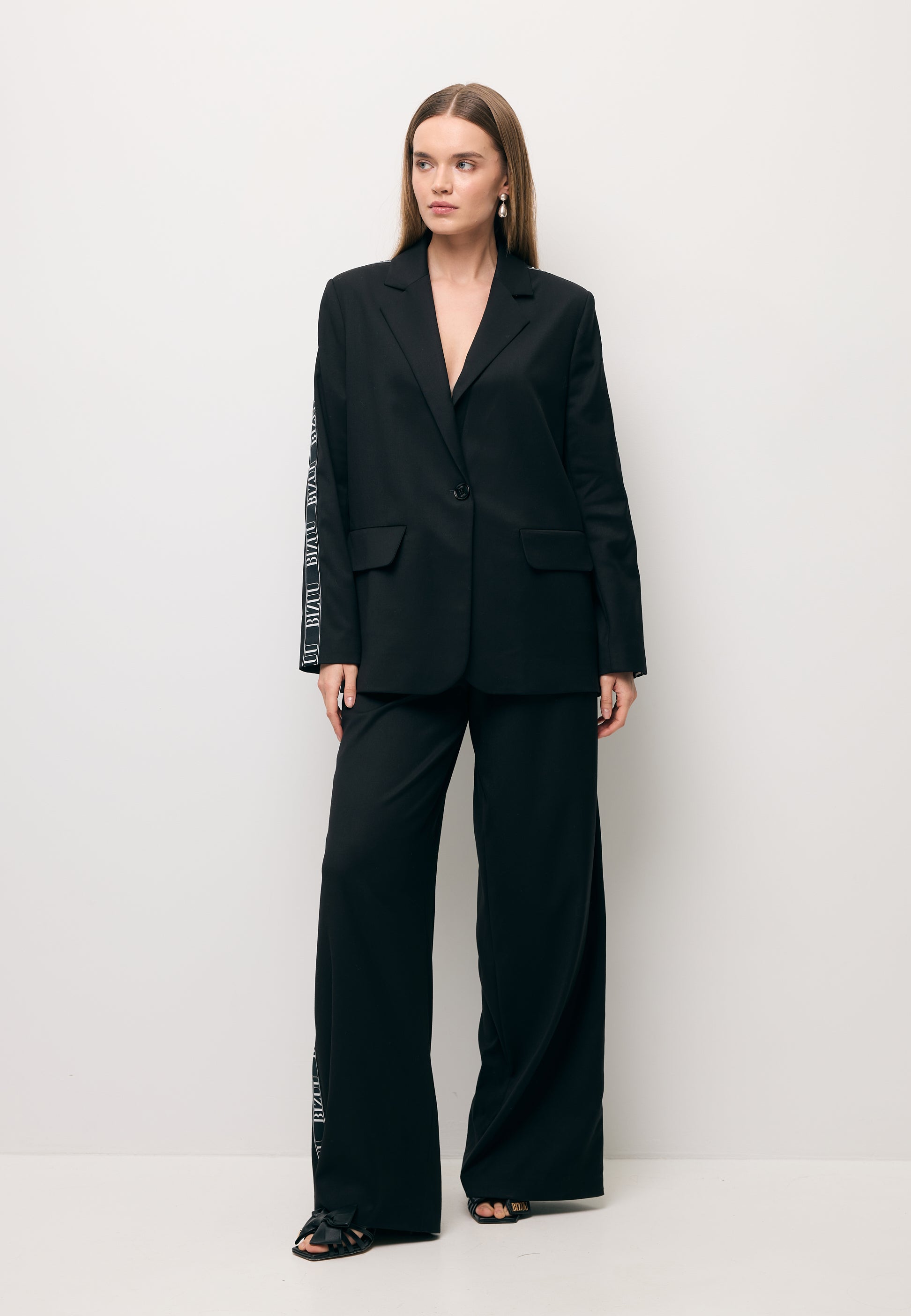 Modelka na tle białej ściany ubrana w czarny garnitur z białym lampasem - marynarka VANDA i spodnie MICHAEL.