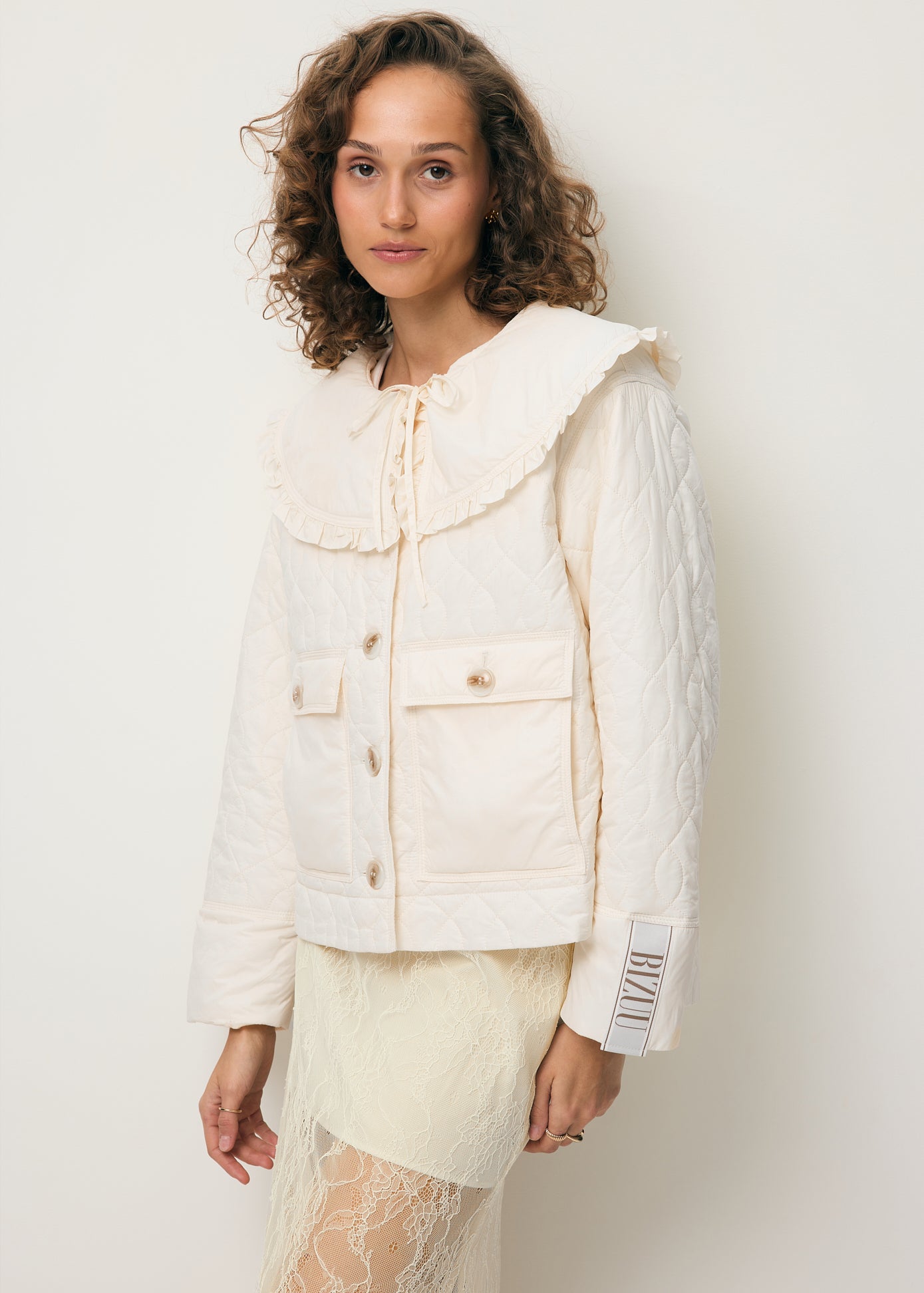 Modelka na tle białej ściany ubrana w kremową kurtkę z kołnierzem ZANNI oraz koronkową spódniczkę FLACE.