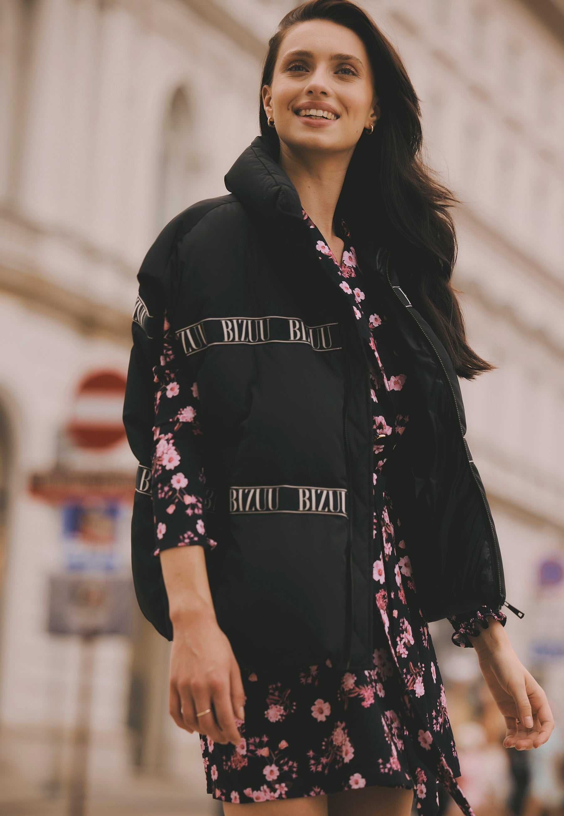 Uśmiechnięta modelka stojąca na ulicy ubrana w czarną puchową kamizelkę AYASE z logowaną taśmą i kwiatową sukienkę GEORGINA.